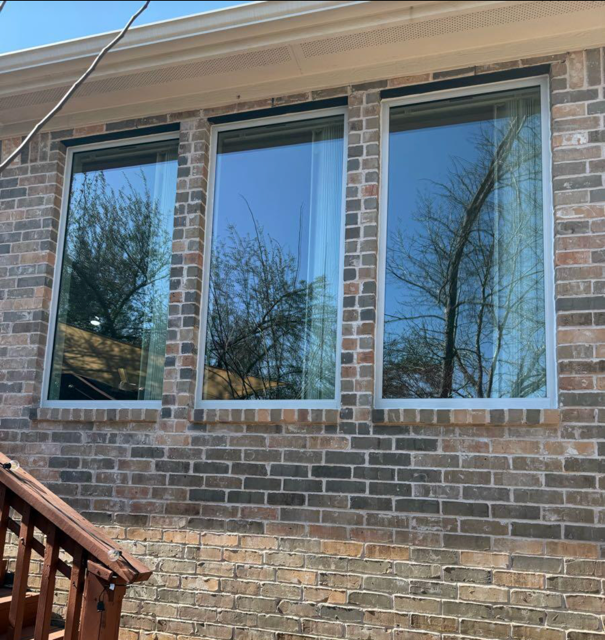 View of a brick house exterior with three large windows reflecting trees and blue sky.