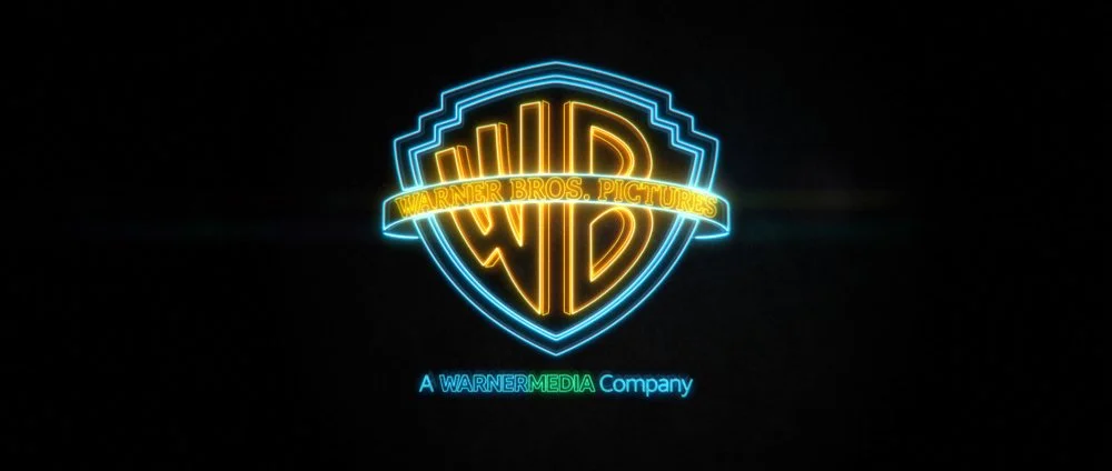 Neon Warner Bros. Pictures logo with blue shield and yellow WB initials, and text 'Warner Bros. Pictures' across the shield and 'A WARNERMEDIA Company' below.