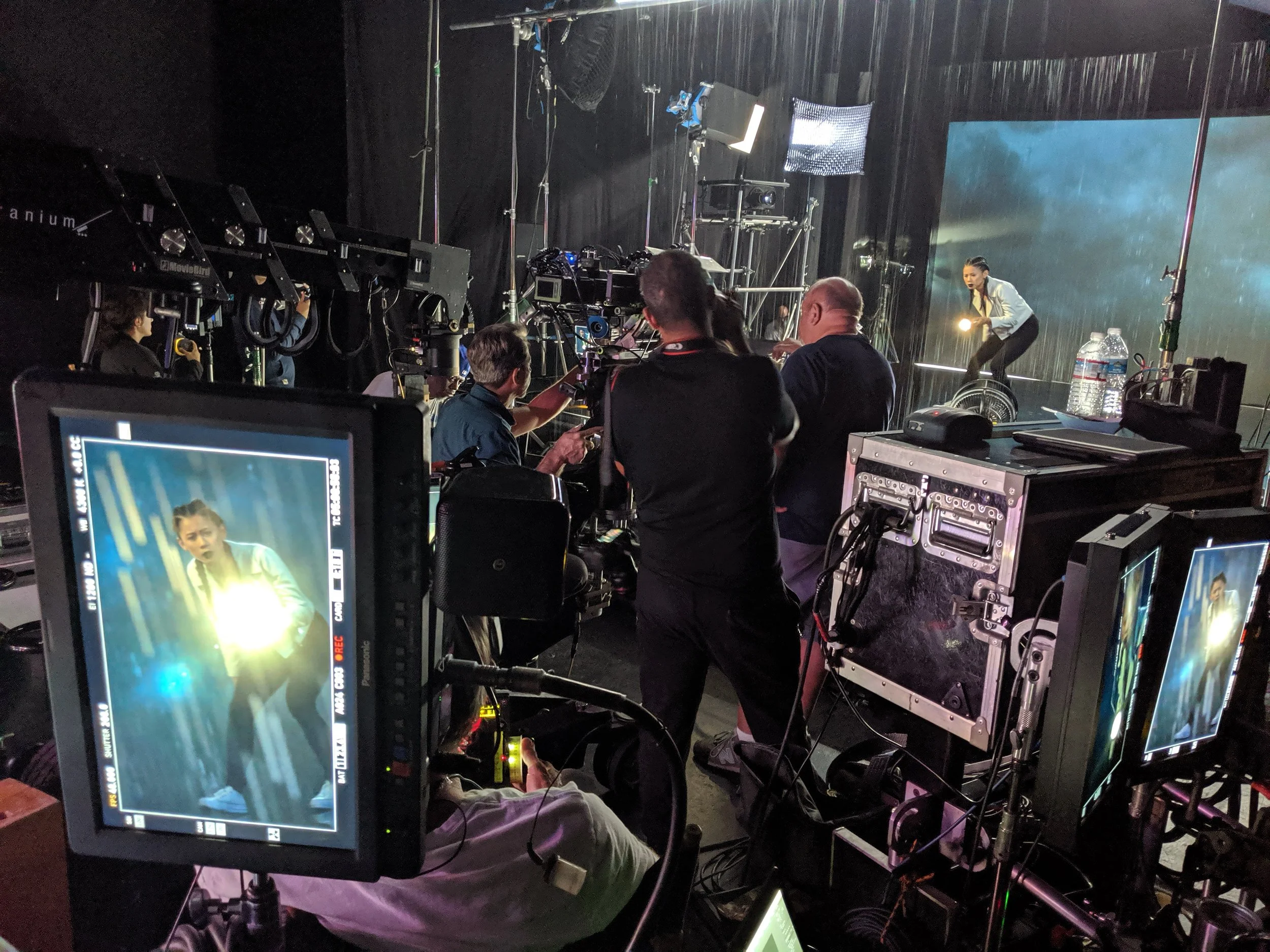 Behind-the-scenes of a video or photo shoot with a woman on a wheeled platform and crew members operating cameras and equipment in a studio. The CW