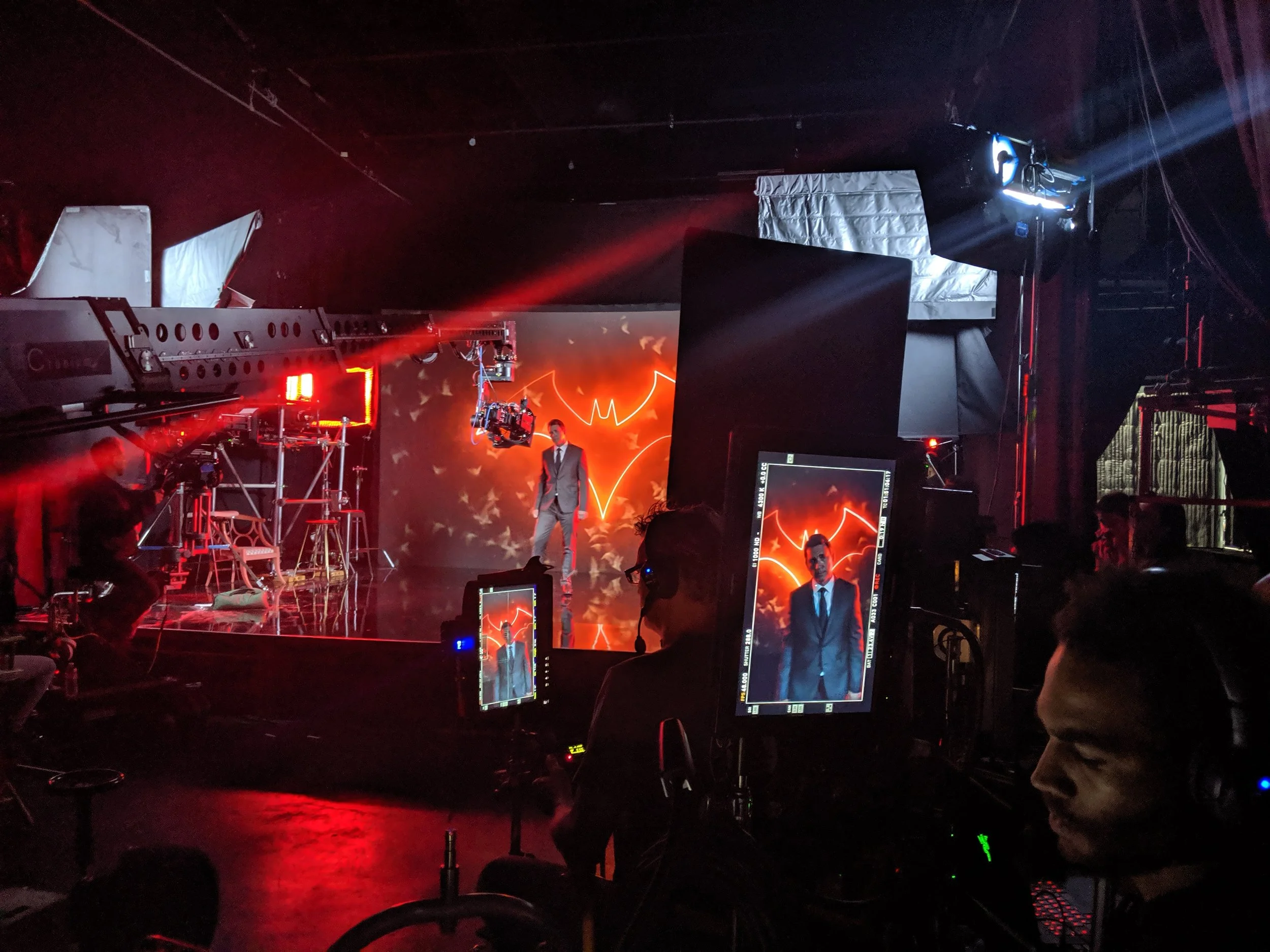 Video production studio with a man in a suit on a stage with a Batman logo behind him, multiple monitors and cameras, crew members working, stage lighting, and a dark environment.