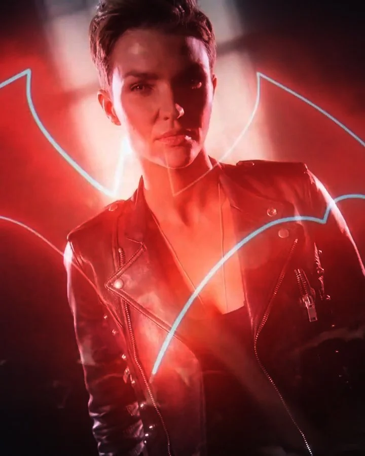 A person with short hair wearing a black leather jacket, standing in front of a glowing red and blue background.