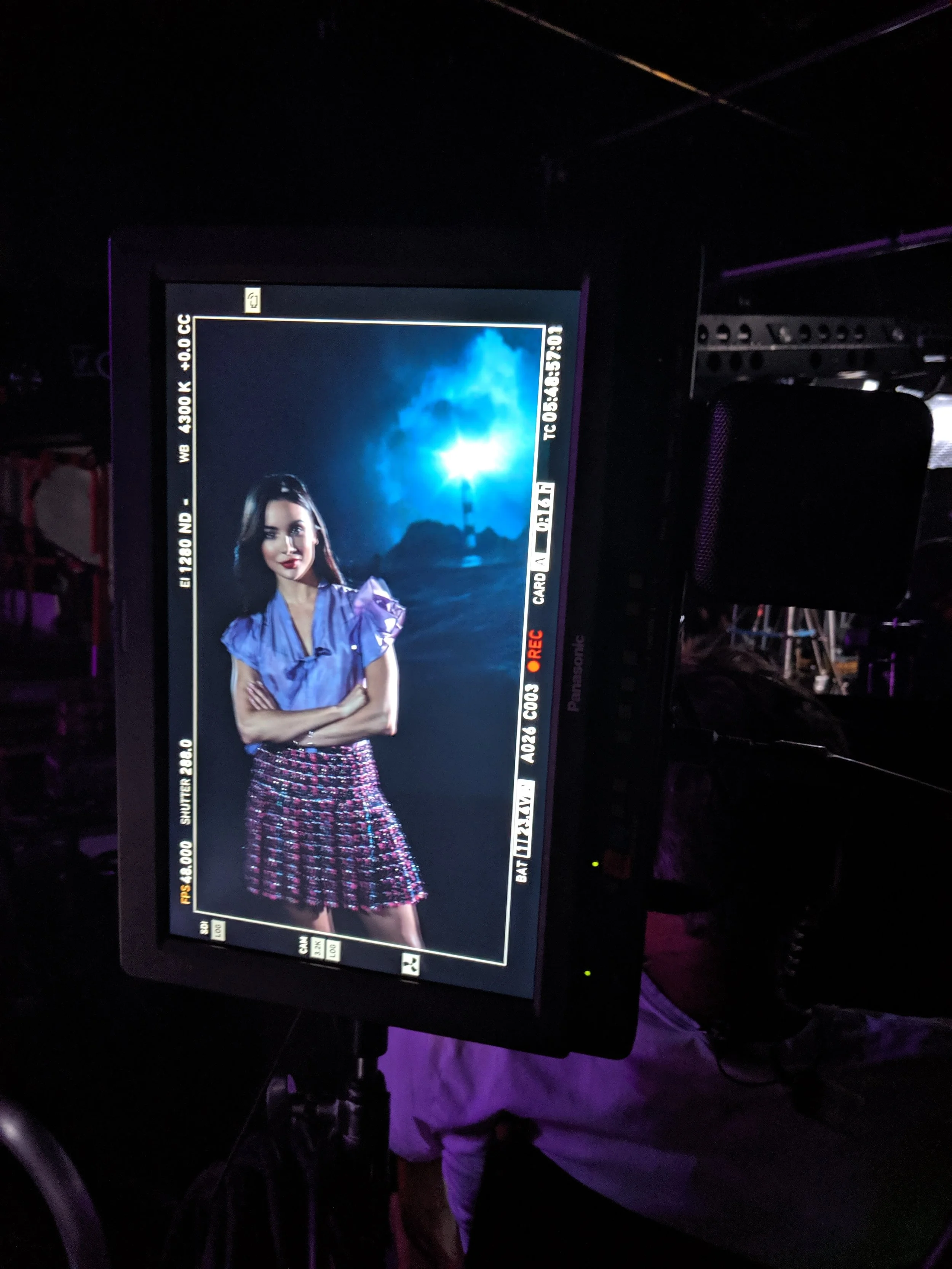 A television screen showing a female model in a purple top and a pink skirt, standing outdoors at night with bright moonlight in the background. The CW