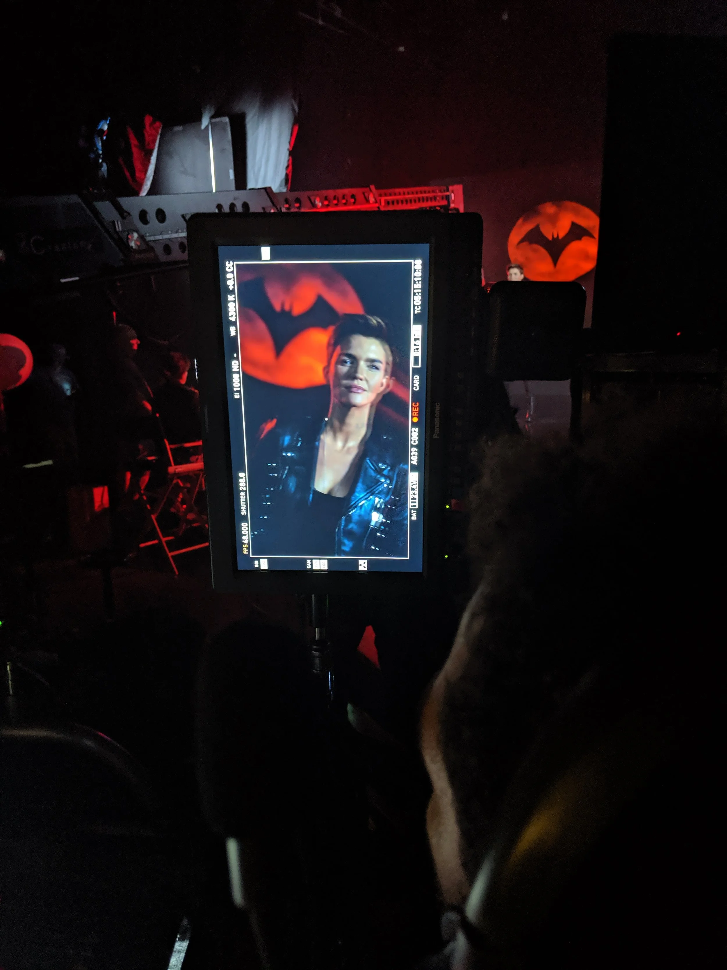 On a film or photo set, a person is looking at a camera monitor that displays a woman with short hair, wearing a black jacket, with a background of a large orange moon featuring a black bat silhouette.