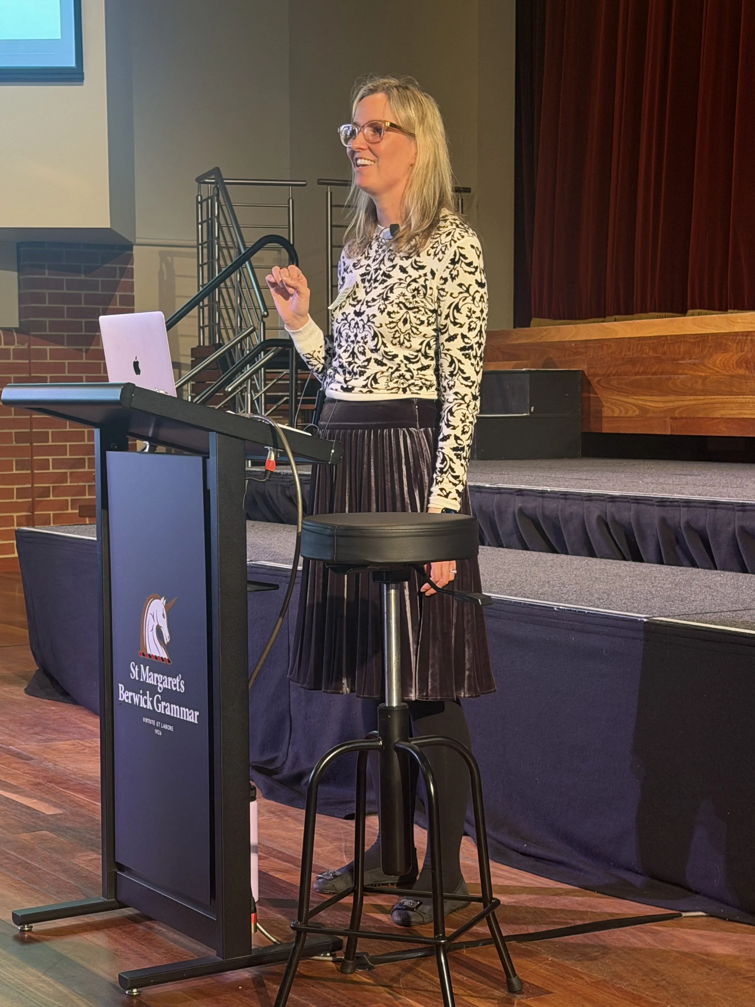 A woman with blonde hair, glasses, and a black and white patterned sweater stands behind a podium with a laptop on it, speaking at a presentation or event with a stage and red curtain in the background.