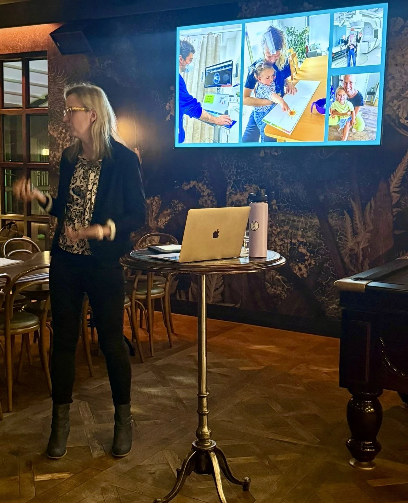 A woman standing in a room with wood flooring, dark patterned wallpaper, and a round table with a laptop and a water bottle, while a large screen displays images of medical and healthcare activities.