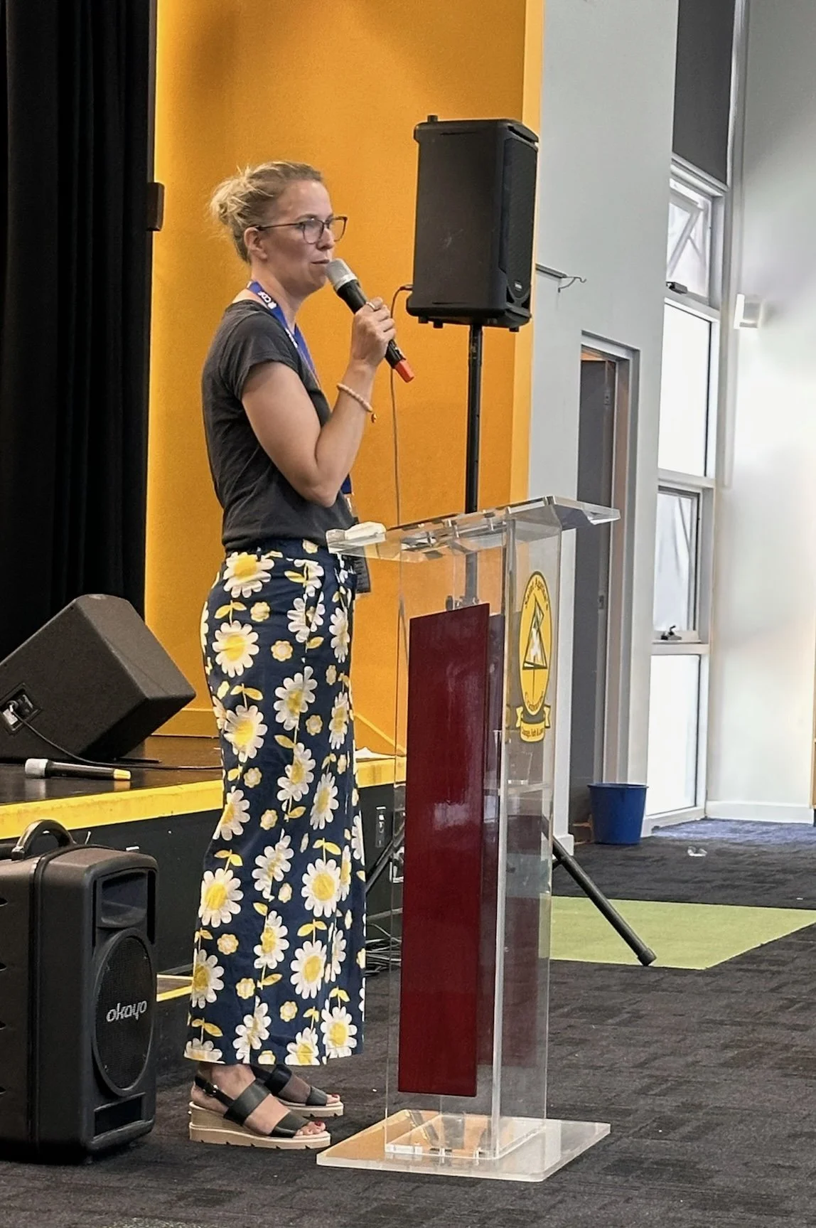A woman wearing glasses, a dark gray t-shirt, and floral pants is standing at a transparent podium speaking into a microphone during a presentation or speech in a room with yellow and gray walls.
