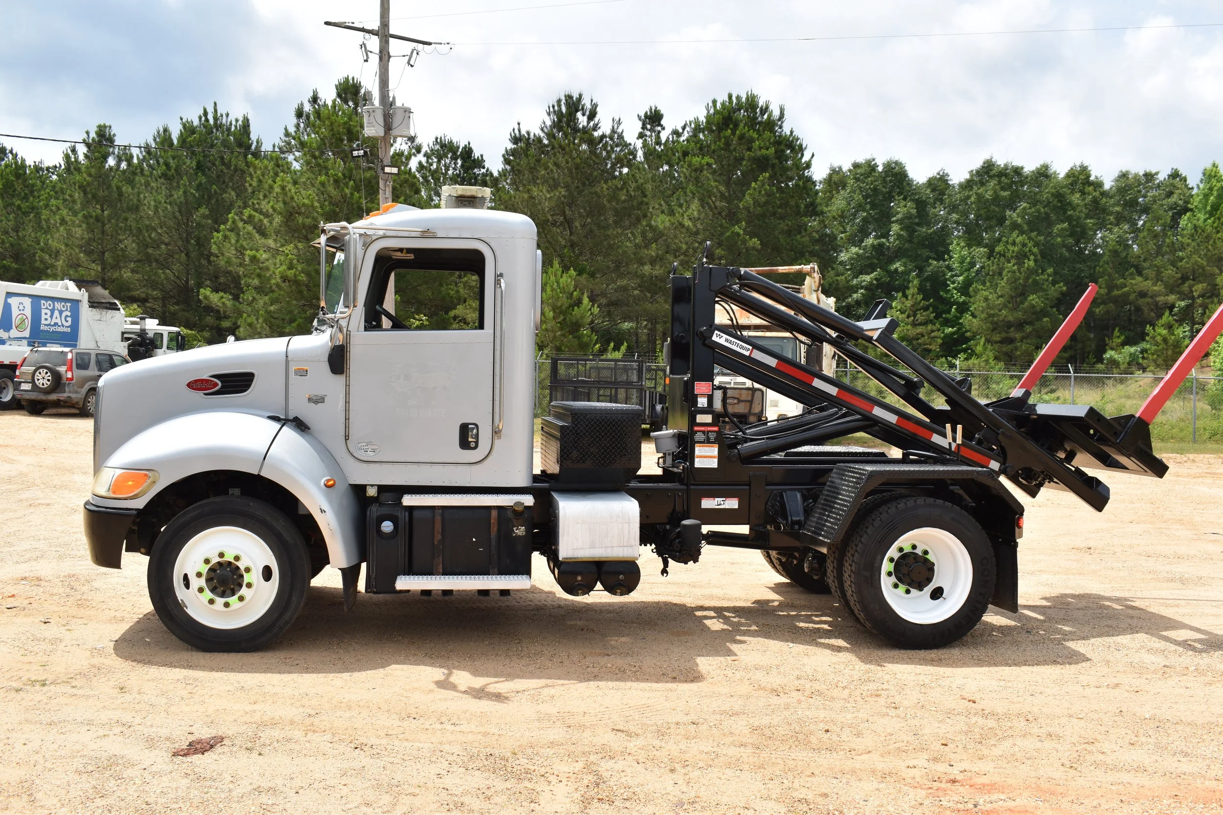 White garbage truck with black mechanical arm on a dirt lot, surrounded by trees and parked vehicles.