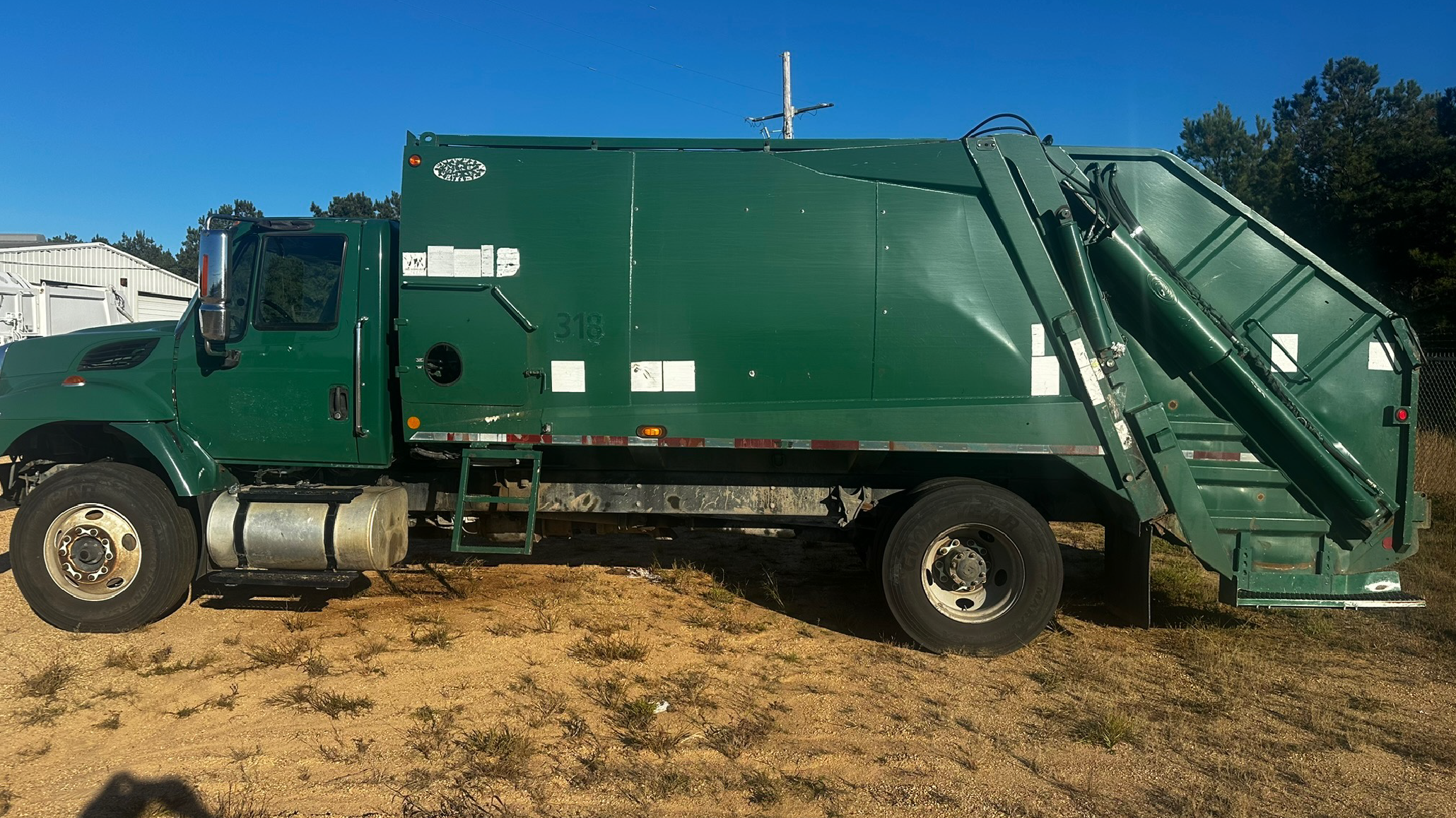 Green garbage truck parked on dirt ground with a clear blue sky in the background.