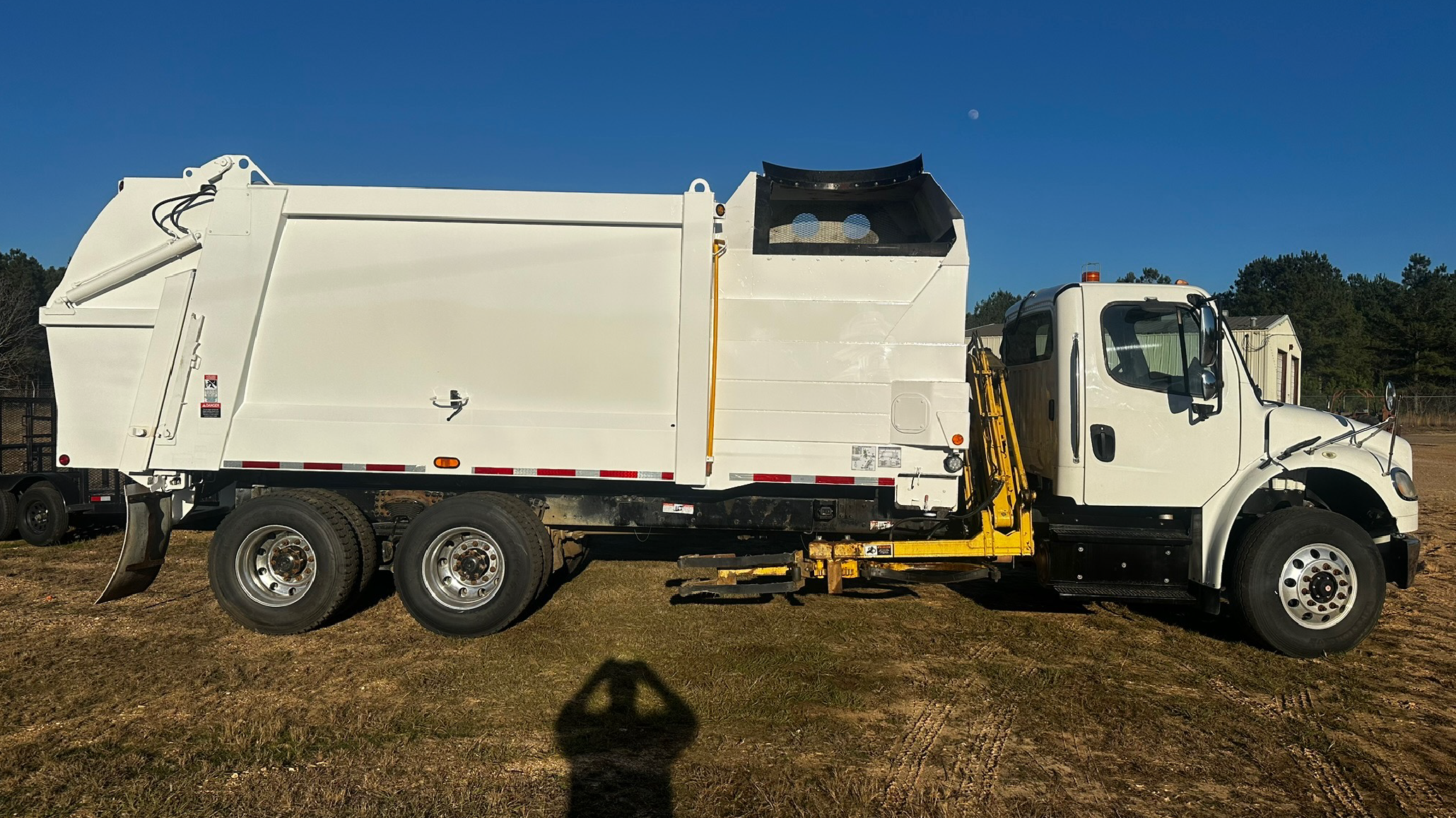 White street sweeper truck parked on dirt ground with clear blue sky and trees in the background.