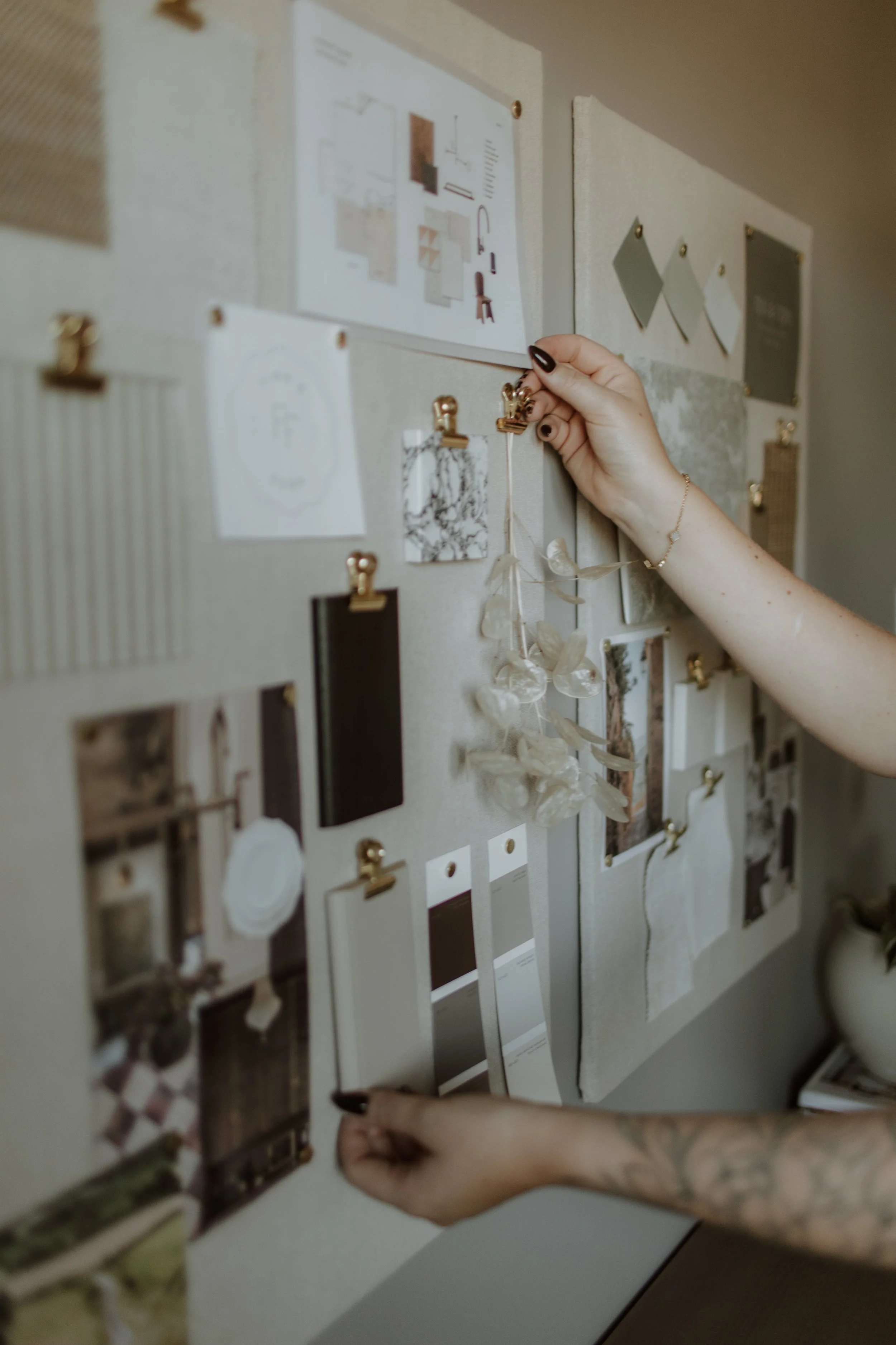 Close-up of two hands pinning photographs and swatches to a gray fabric bulletin board with gold clips, containing images, color samples, and design plans.