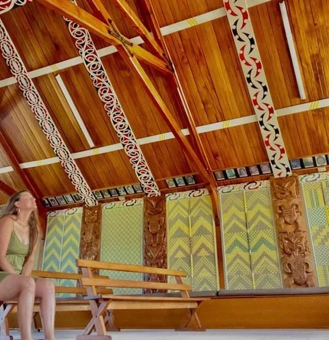 A woman sitting on a bench inside a traditional Polynesian building with carved wooden pillars and colorful patterned walls.