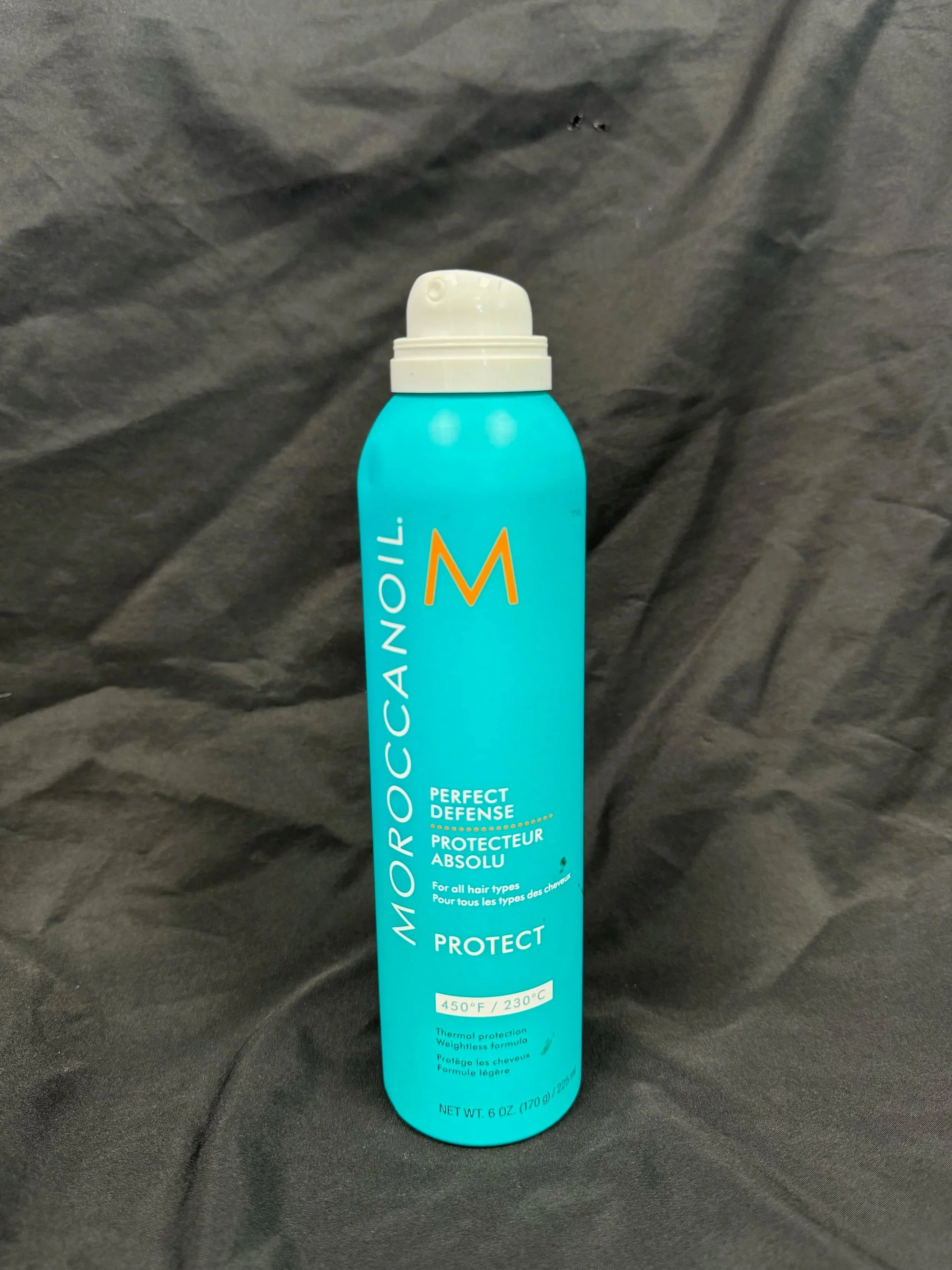 A tall turquoise bottle of Moroccanoil Perfect Defense heat protectant spray with a white spray cap, placed against a dark fabric background.
