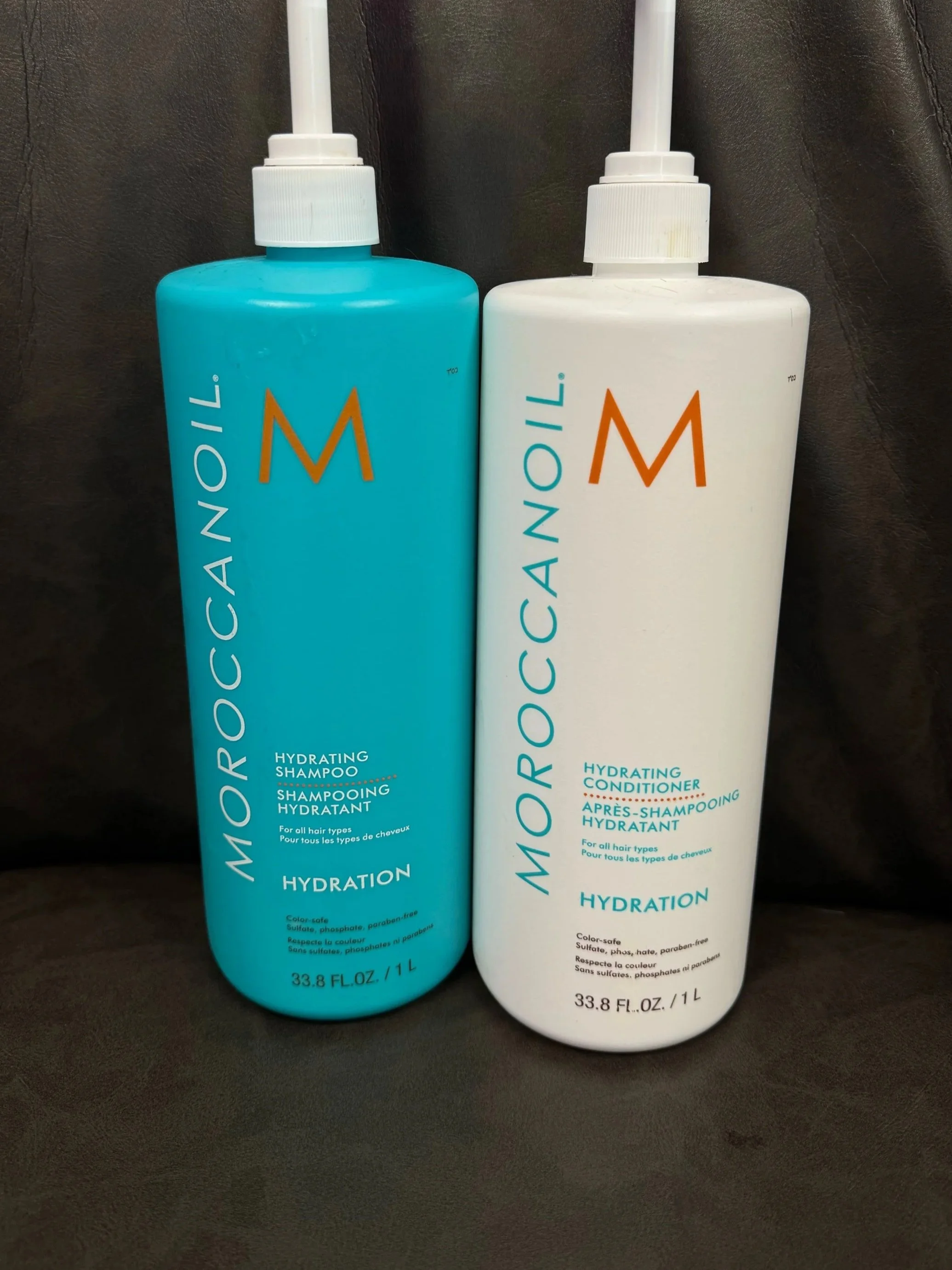Two bottles of Moroccanoil hair products, one blue hydrating shampoo and one white hydrating conditioner, standing upright on a dark surface.