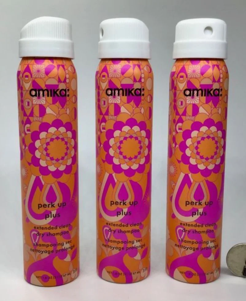 Three bottles of Amika perk up plus extended clean dry shampoo for extending hair freshness, with a colorful pink, orange, and purple floral pattern on the bottles, placed next to a coin for scale.