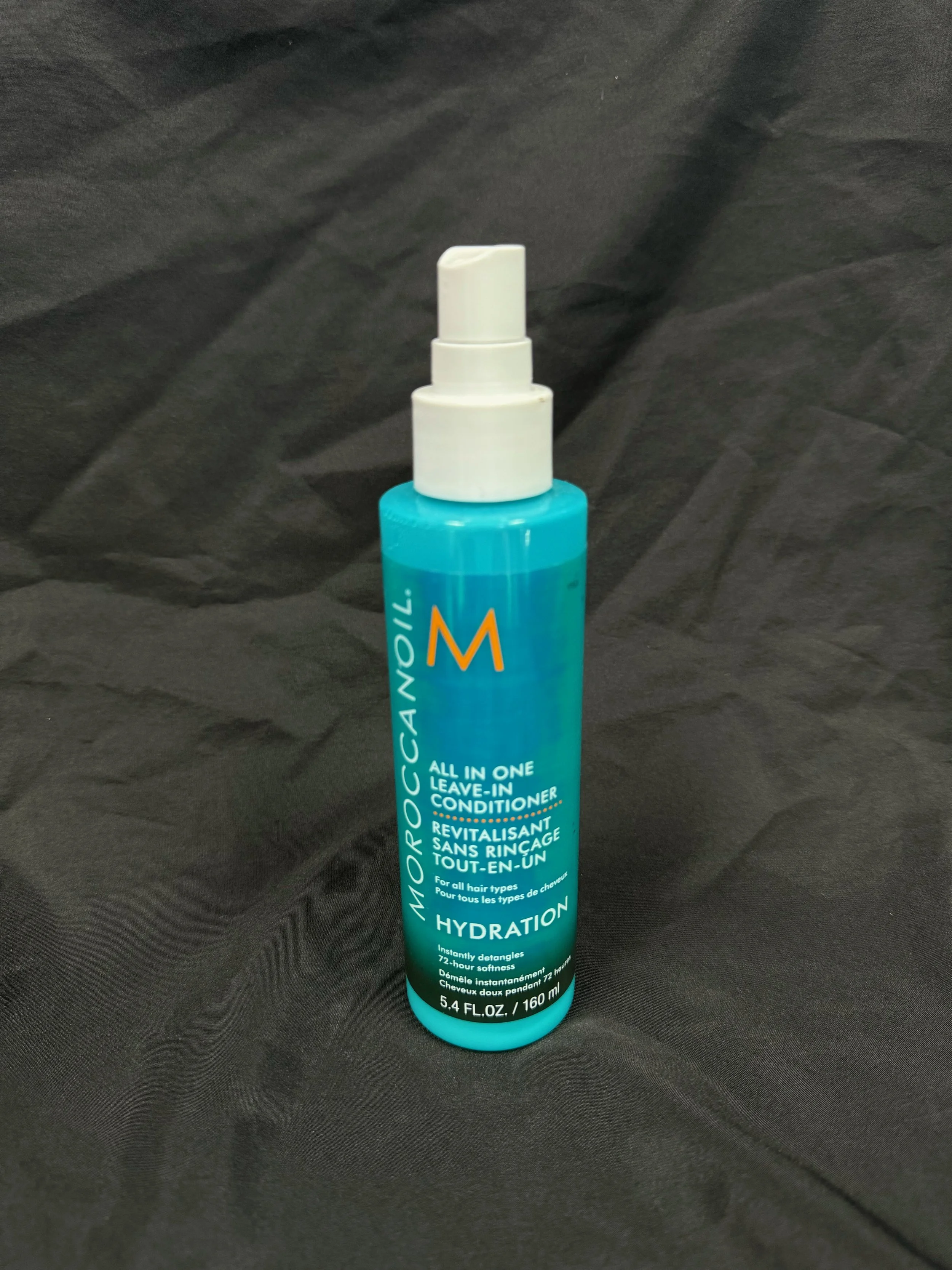 A blue bottle of Moroccanoil All-in-One Leave-in Conditioner with white pump dispenser, placed on black fabric background.