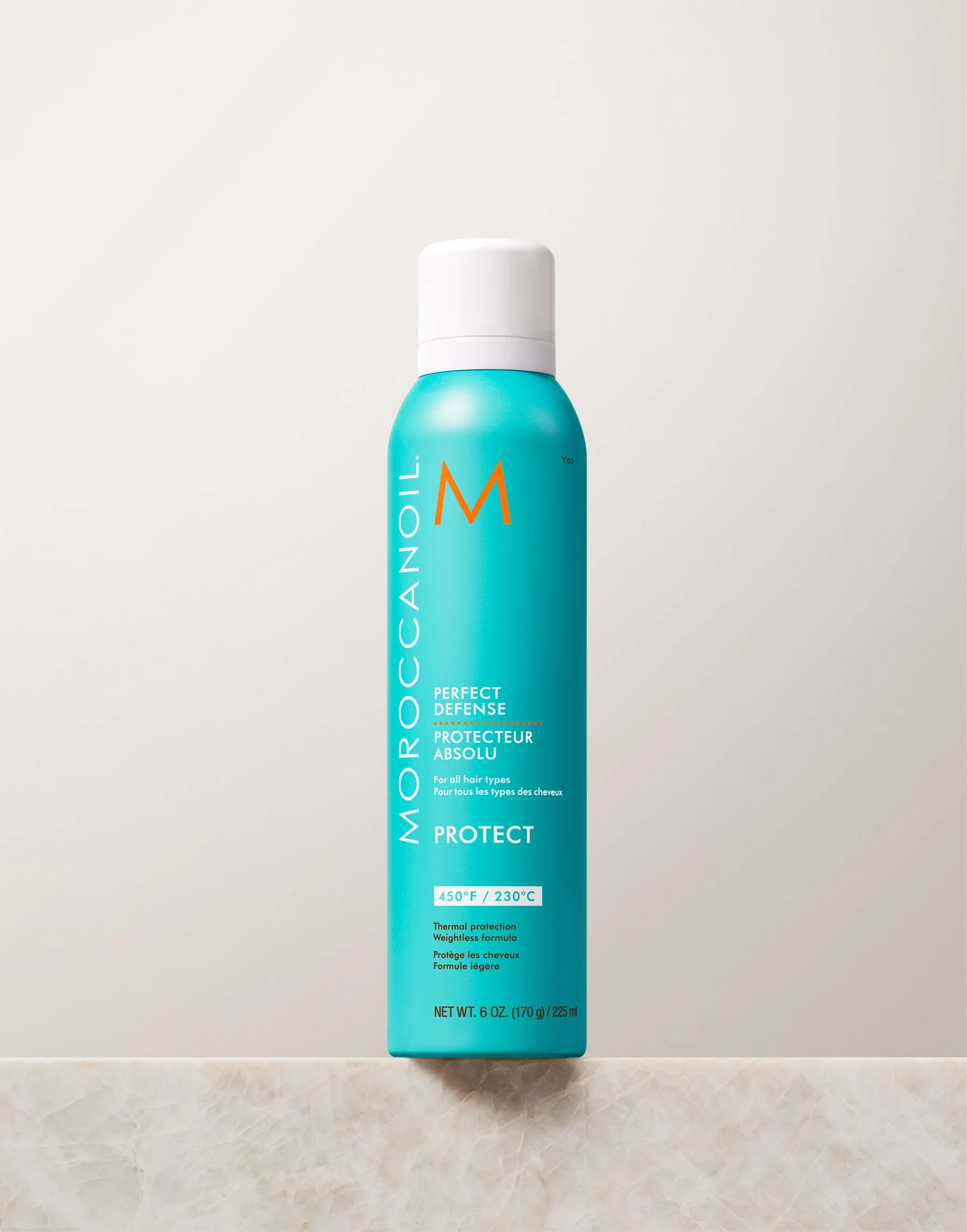 A tall turquoise bottle of Moroccanoil Perfect Defense heat protectant spray with a white spray cap, placed against a dark fabric background.