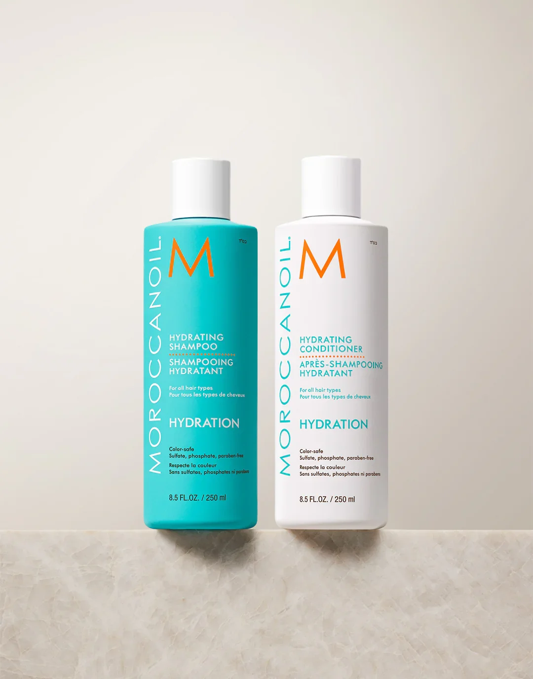 Two bottles of Moroccanoil hair products, one blue hydrating shampoo and one white hydrating conditioner, standing upright on a dark surface.