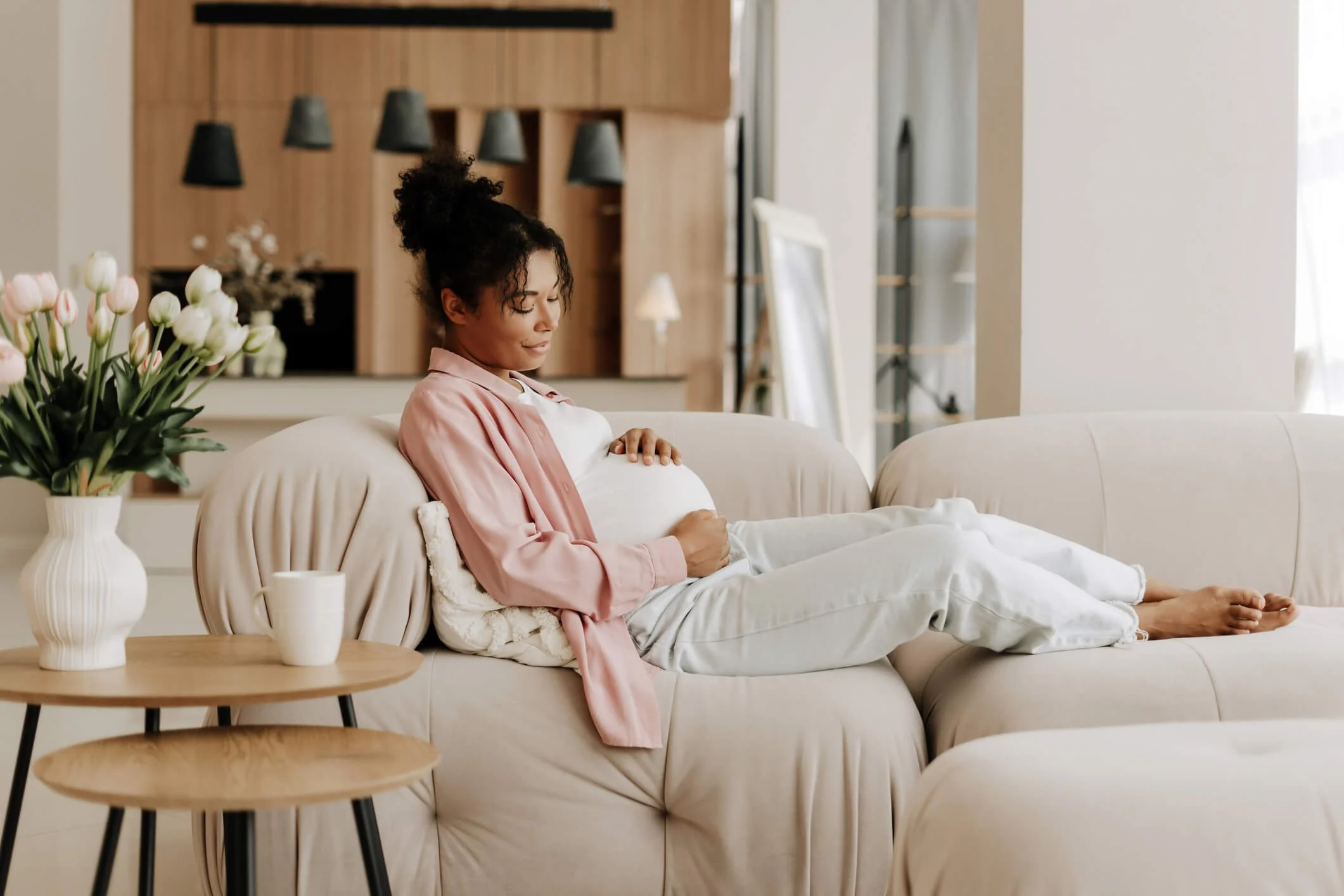 A woman sitting on a beige couch, holding her pregnant belly, in a bright living room with modern decor, a side table with a vase of pink tulips, and natural light coming from a window.