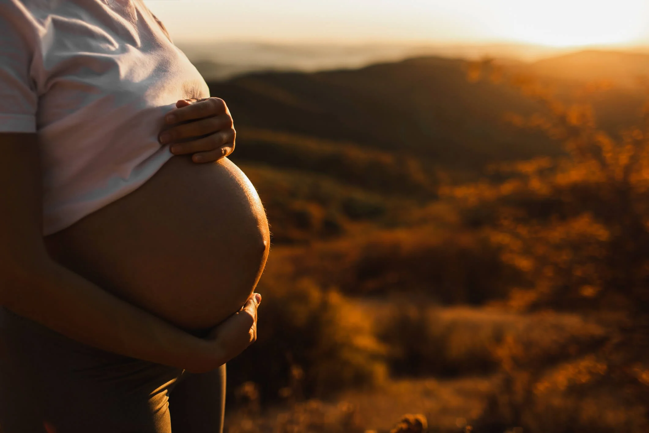 A pregnant woman holding her belly with one hand, wearing a white shirt, standing outdoors during sunrise, with a landscape of hills in the background.