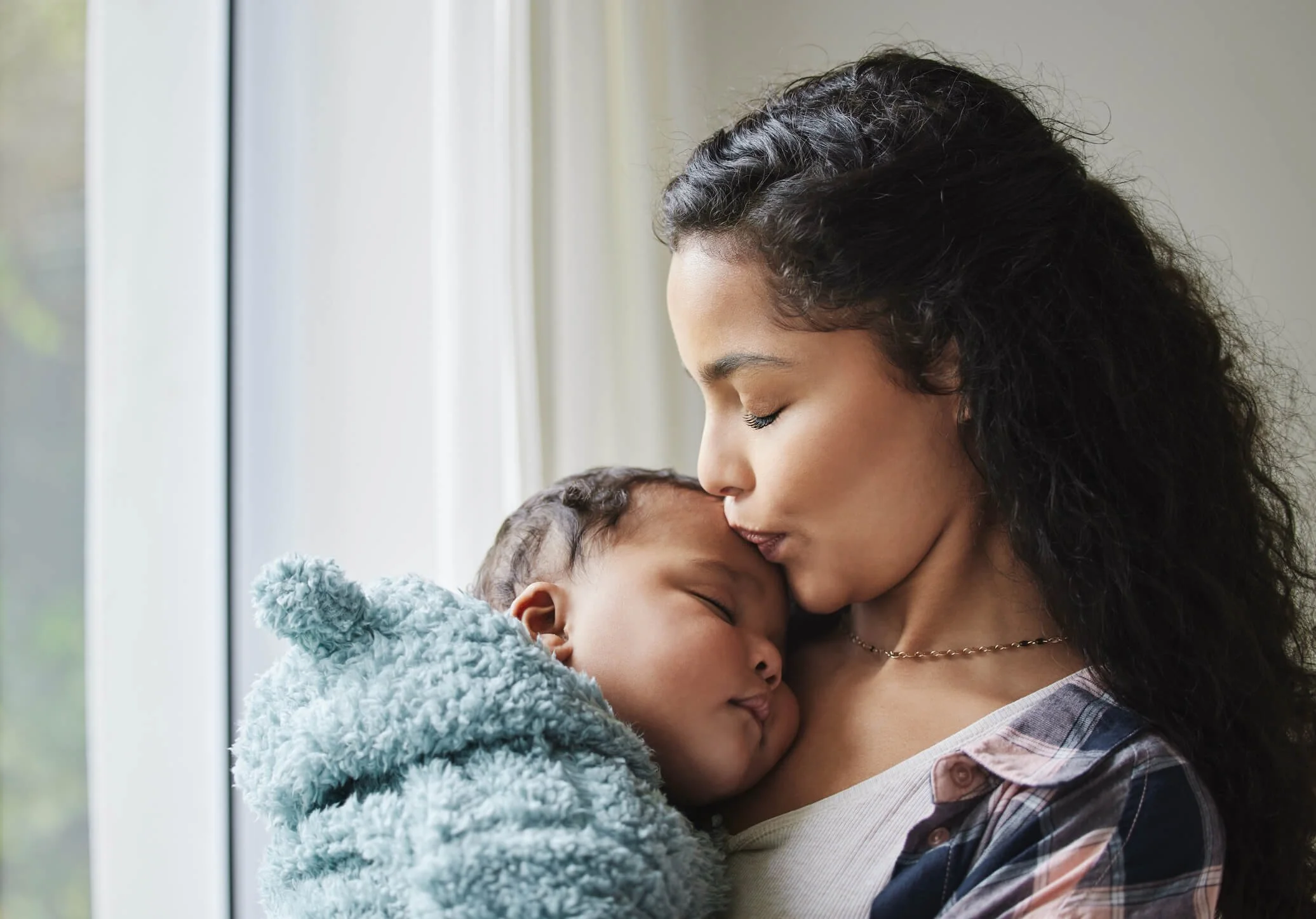 A woman with curly hair gently kisses a sleeping baby on the forehead. The baby is wrapped in a soft, furry gray blanket. They are near a window with natural light, creating a warm, tender moment.