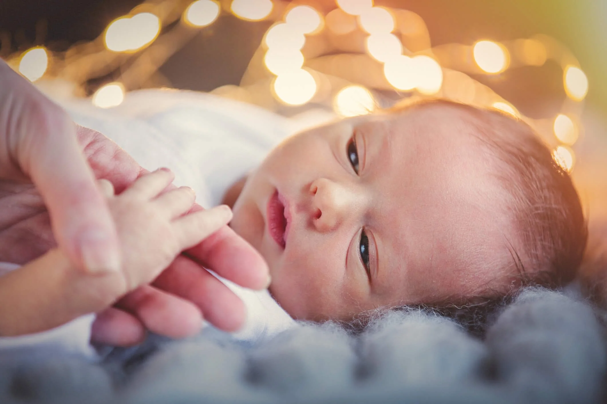 Close-up of a newborn baby lying on a soft surface, looking at an adult's hand touching their chin with warm, glowing string lights in the background.