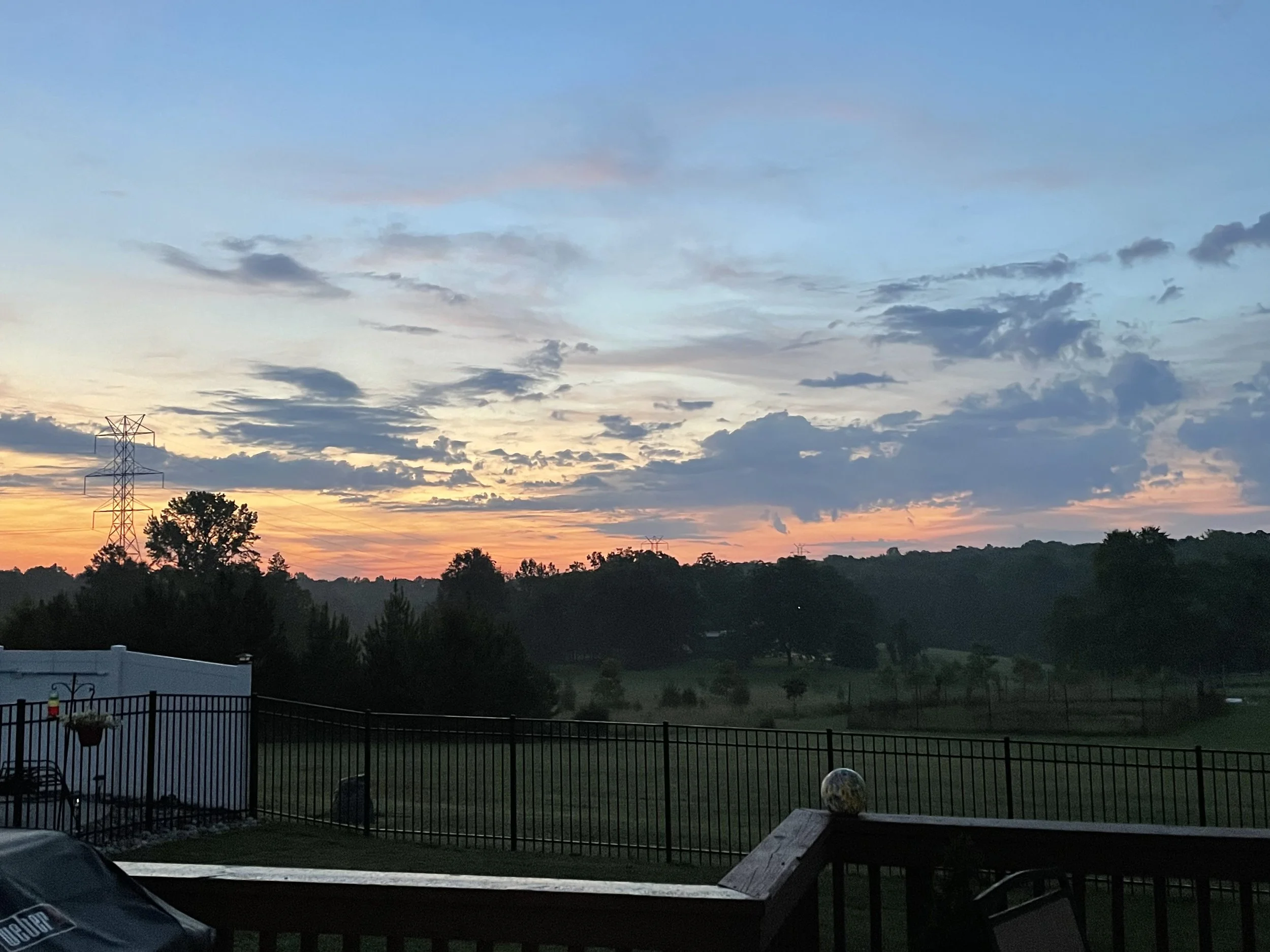 Sunrise sky with scattered clouds over a landscape of trees, hills, and a fenced backyard with outdoor furniture.
