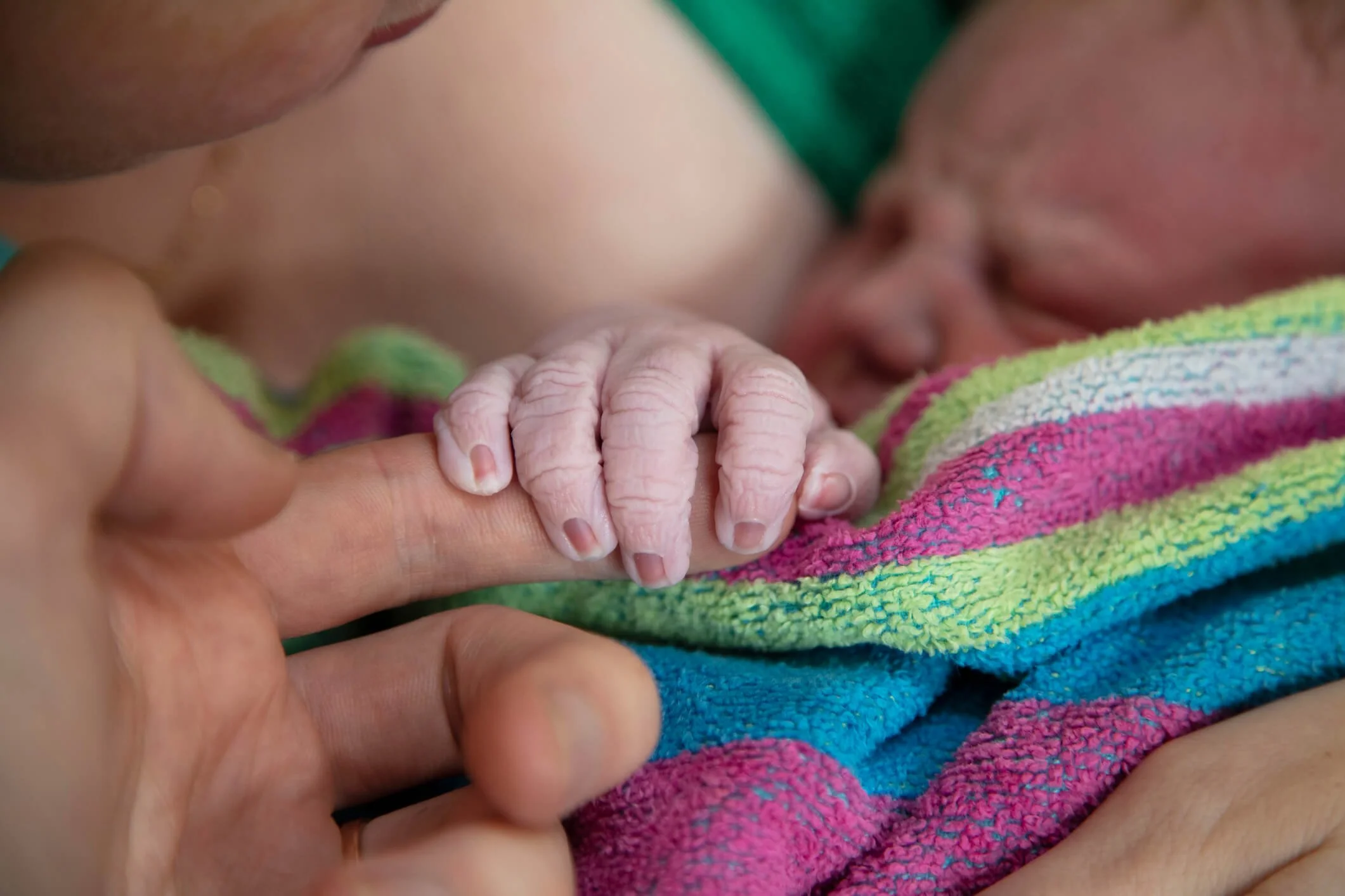 An adult holding a newborn baby's hand, with the newborn wrapped in a colorful striped blanket.
