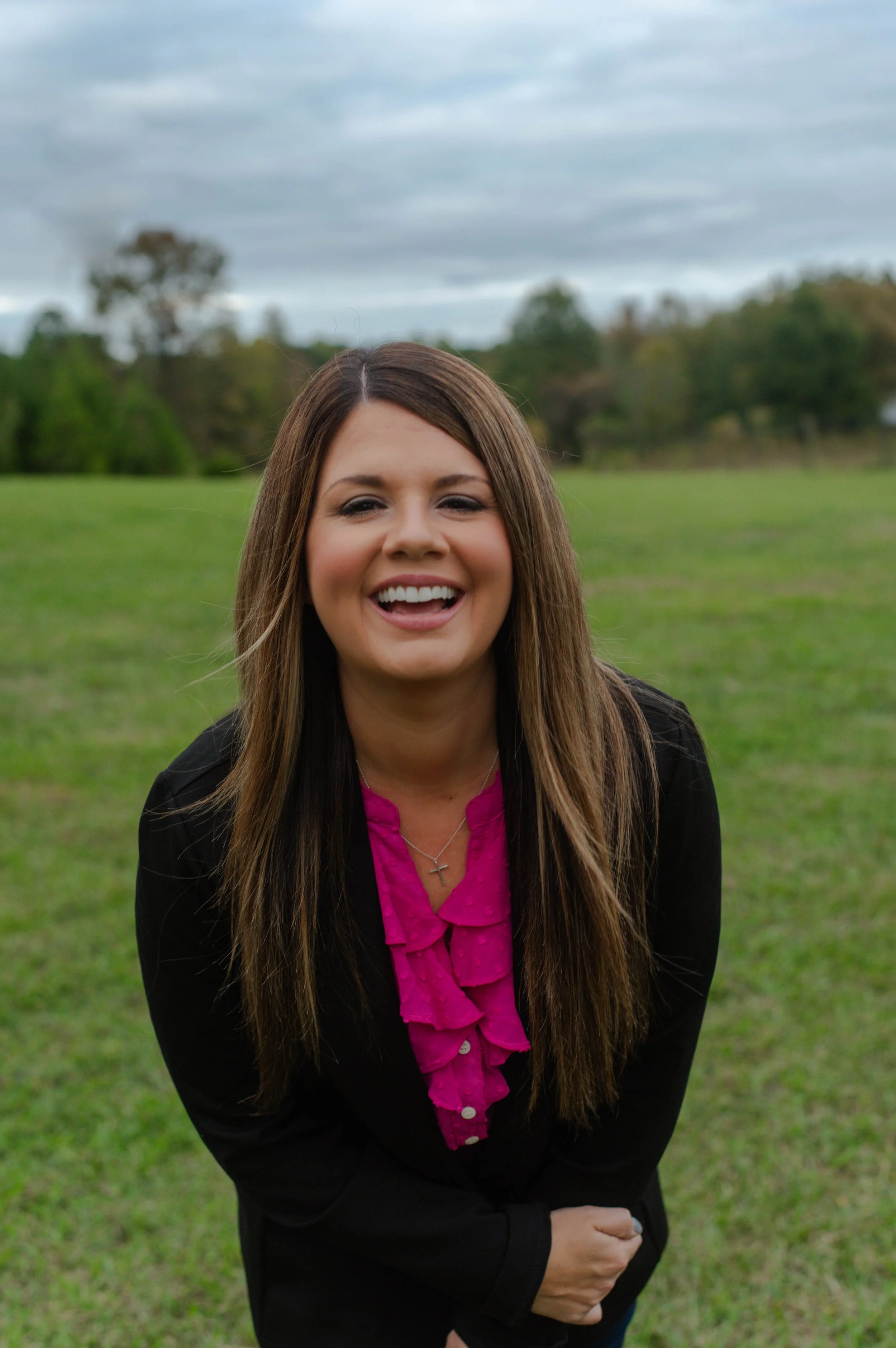 A woman with long brown hair, smiling, standing outdoors on a grassy field with trees and a cloudy sky in the background.