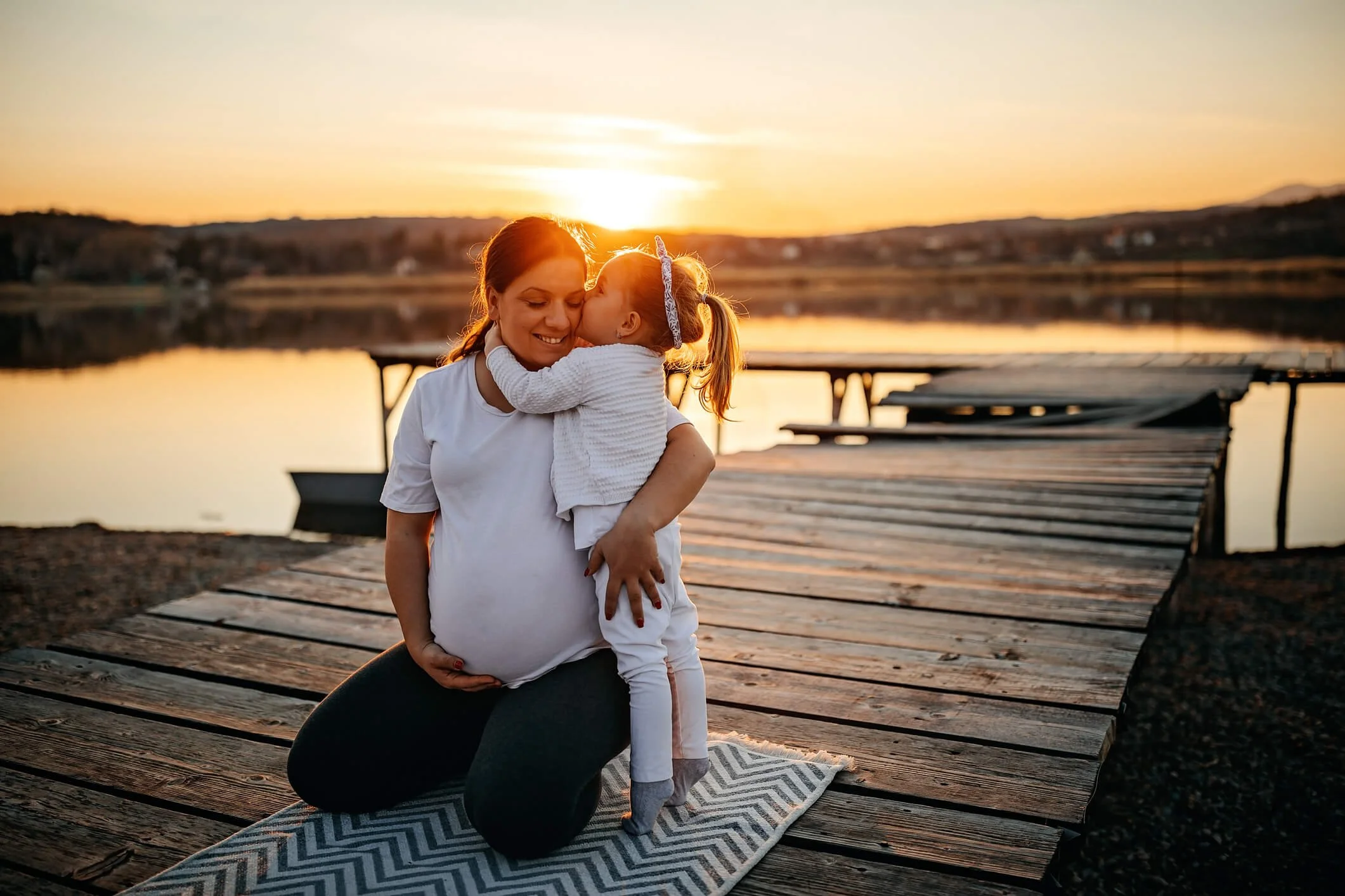 A woman kneeling on a blanket on a wooden dock by the water, holding a young girl, likely her daughter, who is giving her a kiss on the face. The sunset casting a warm glow in the background.