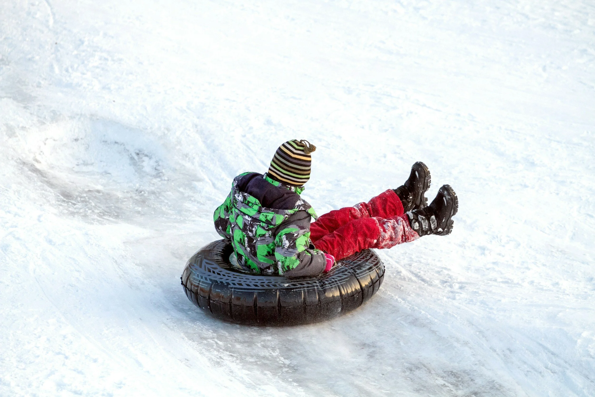 Child in winter clothing riding a snow tube on snow-covered ground.