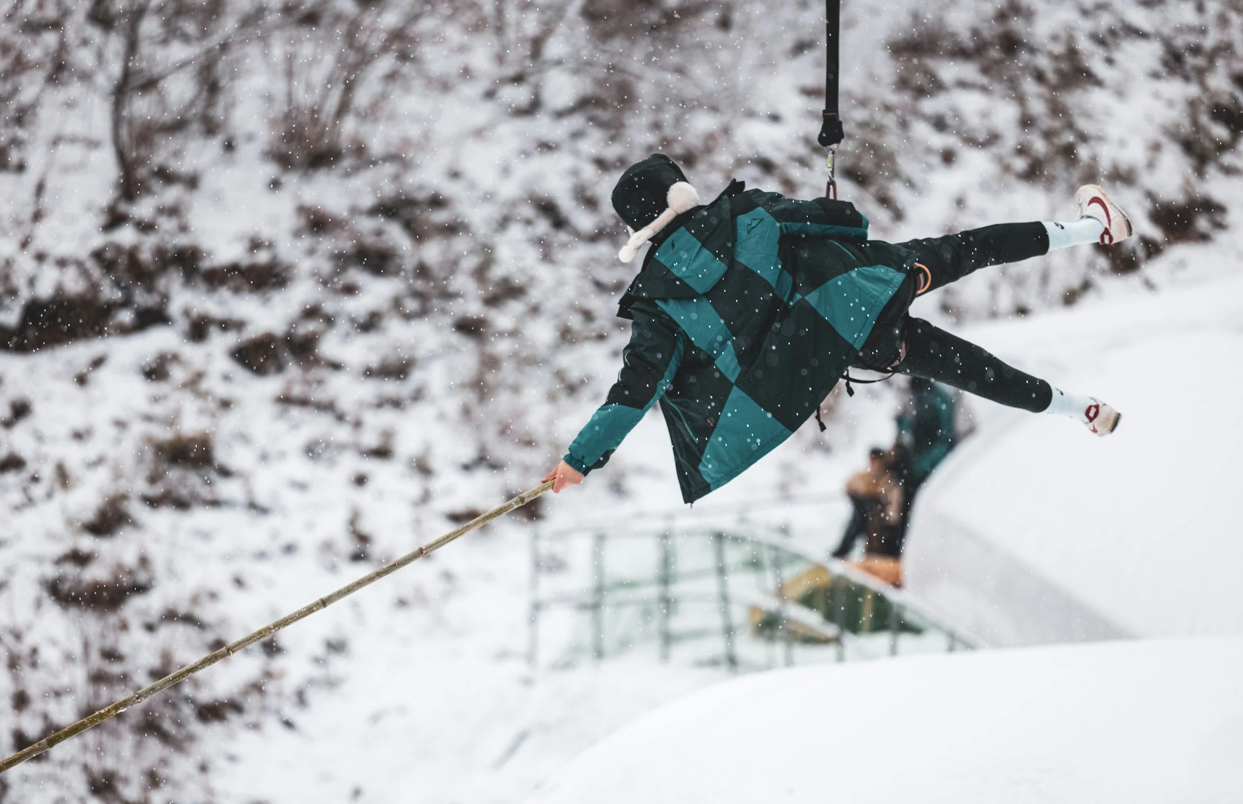 A person in a blue and black winter coat and a black hat is flying upside down on a harness over a snowy landscape, holding onto a rope with one hand.