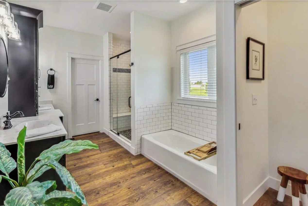 Bright bathroom with white subway tile walls, a bathtub beneath a window with blinds, a glass-enclosed shower with white tile and a dark accent stripe, a wooden floor, a farmhouse-style sink with a black cabinet, and a small plant in the foreground.