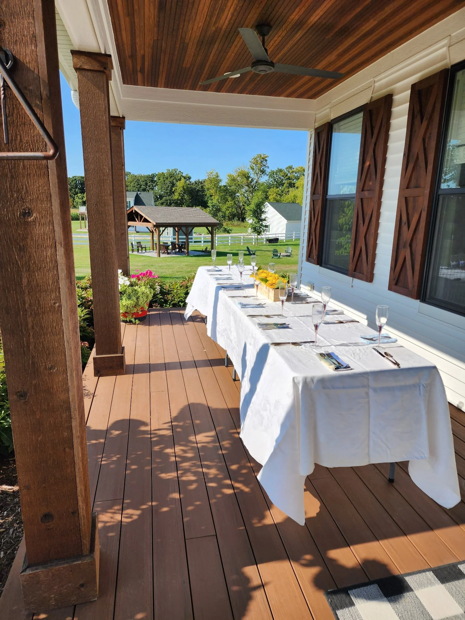 Outdoor porch with a set dining table covered with a white tablecloth, set with utensils and glasses, overlooking a grassy yard with trees, a gazebo, and a small white building in the background.