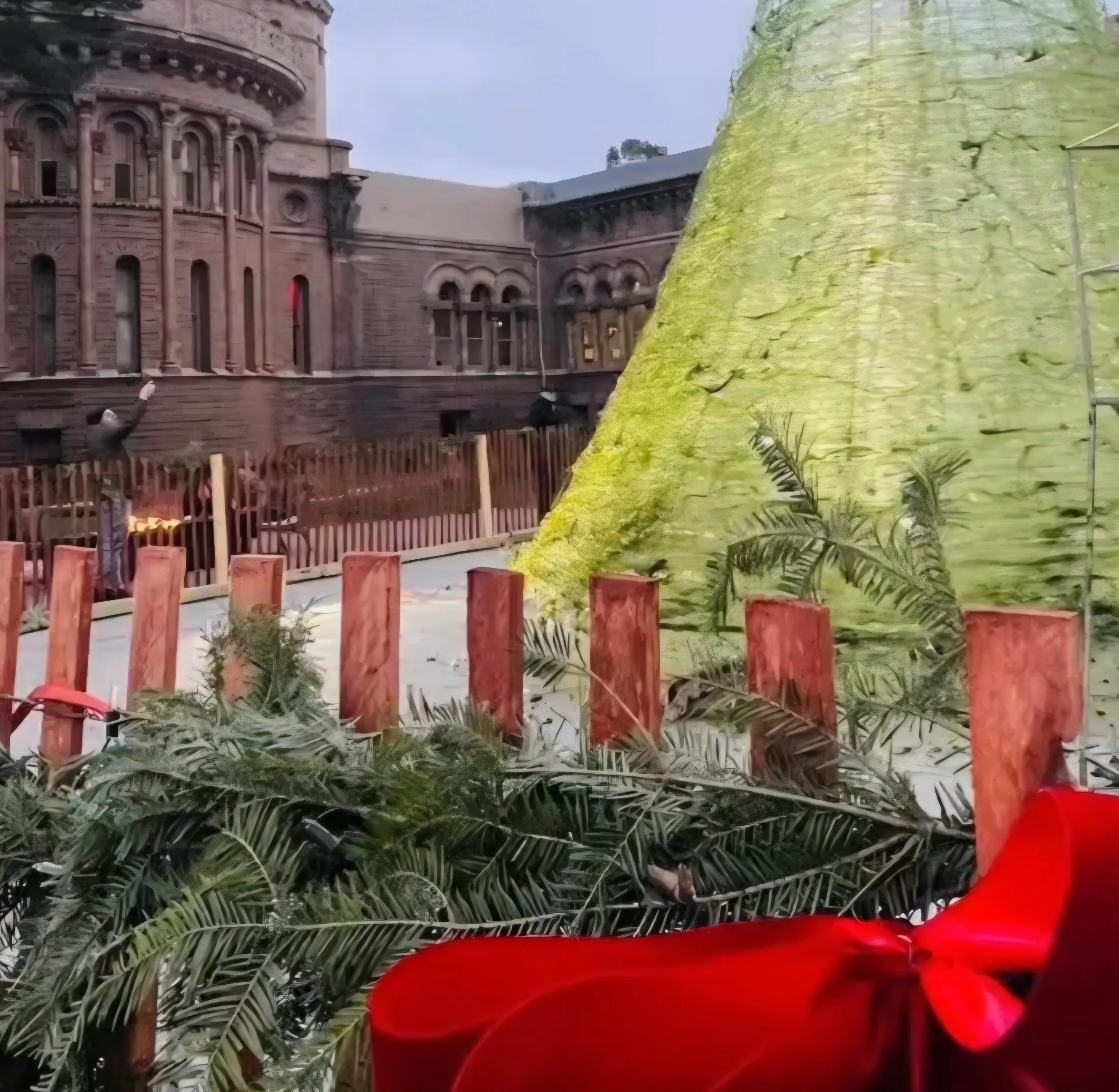 A Christmas tree decorated with a red bow, surrounded by pine branches, in front of a cityscape with historic buildings, a wooden fence, and a pinkish-brown castle-like structure.