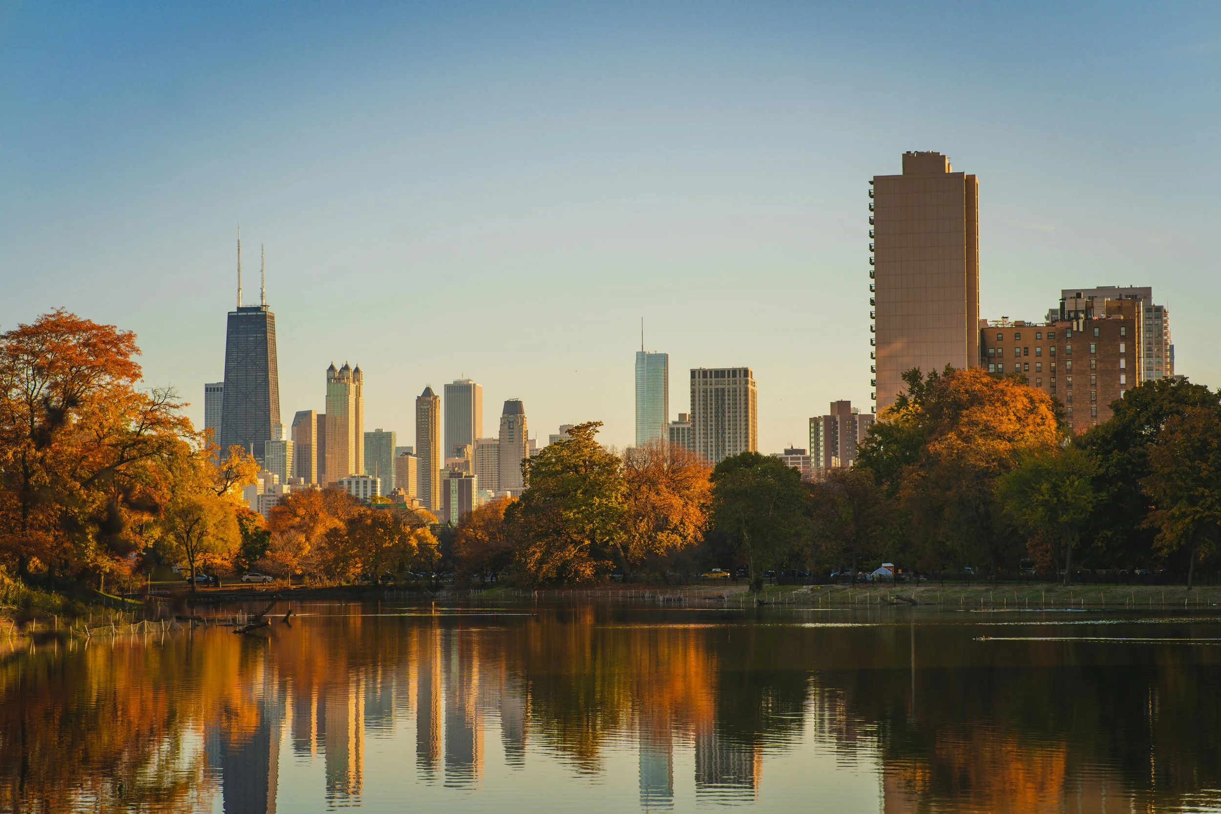 City skyline with tall buildings and trees with autumn-colored leaves reflected in a body of water.