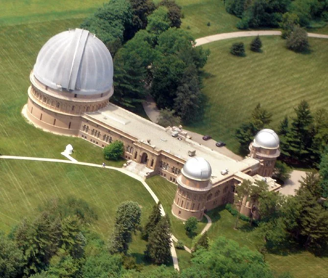 Aerial view of the Griffith Observatory with a large dome and two smaller domes, surrounded by green trees and a park.
