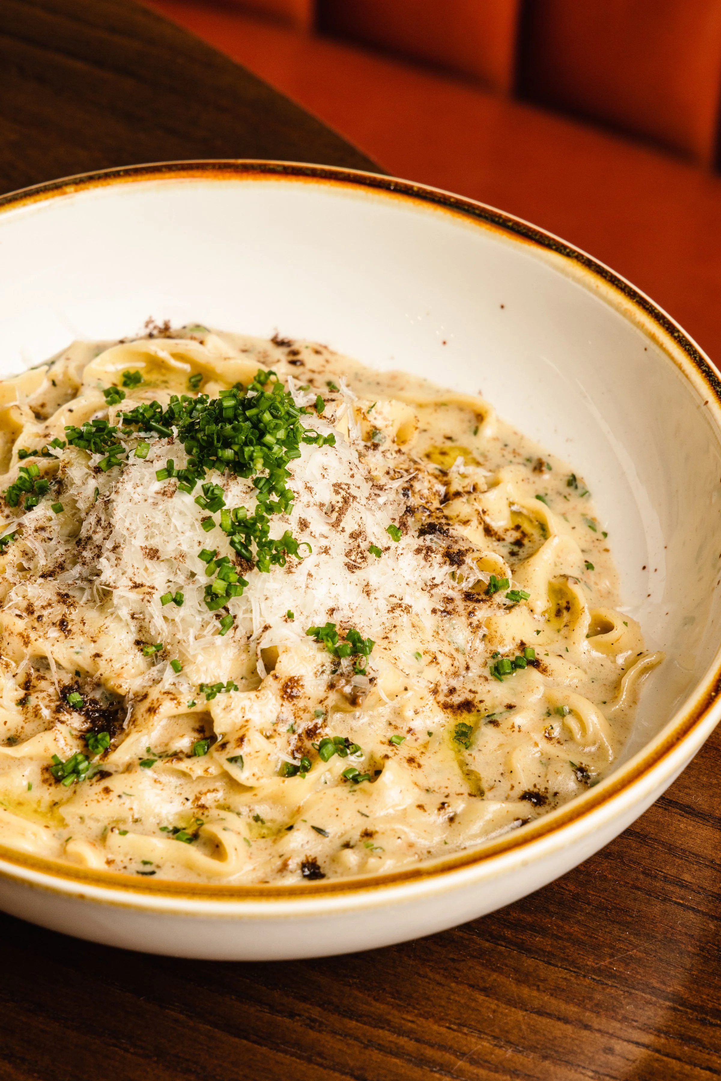 A bowl of creamy Alfredo pasta topped with chopped chives and grated cheese, served on a wooden table.