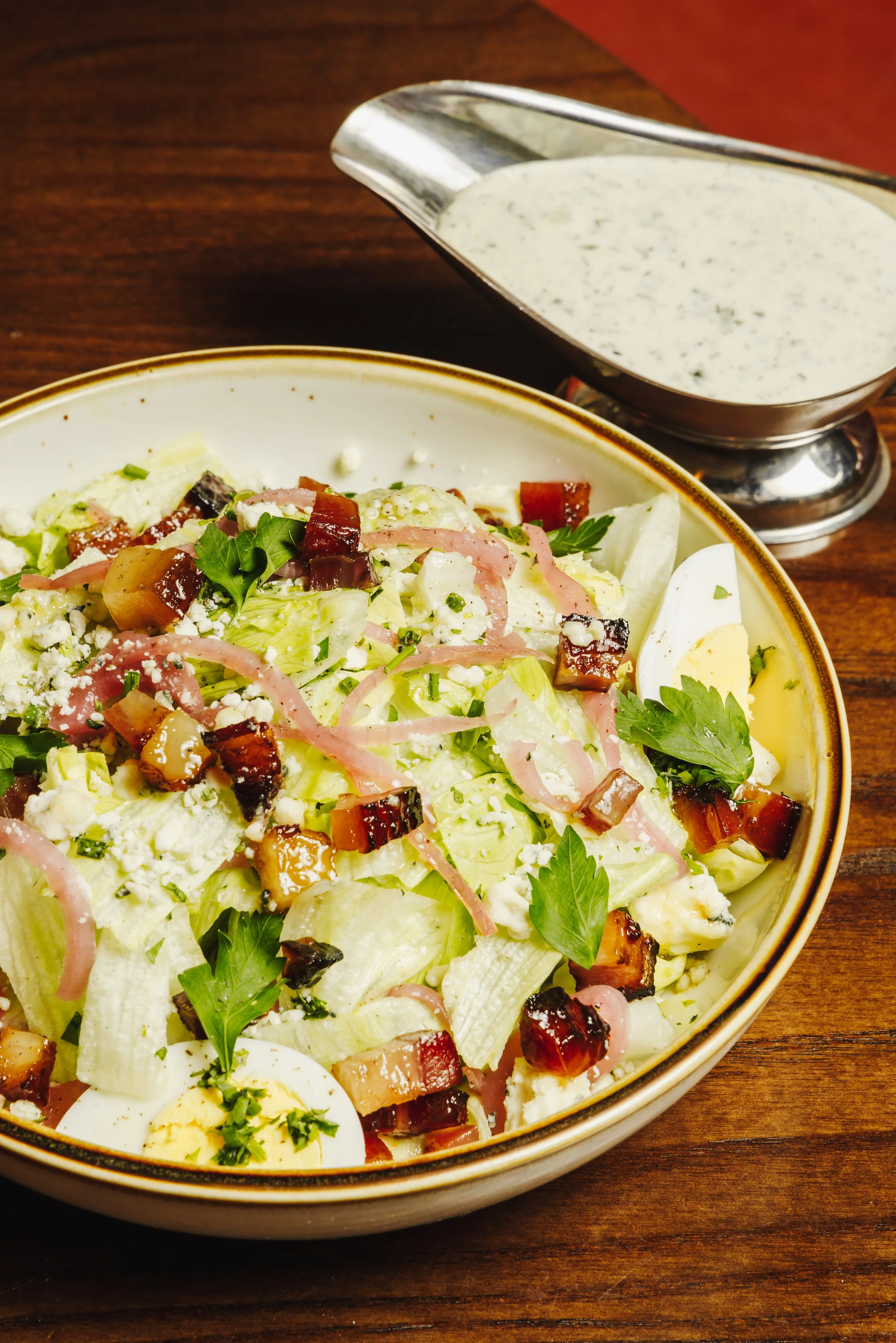 Bowl of chopped iceberg lettuce salad with bacon bits, sliced egg, herbs, and cheese, with a side of creamy dressing in a metal gravy boat.