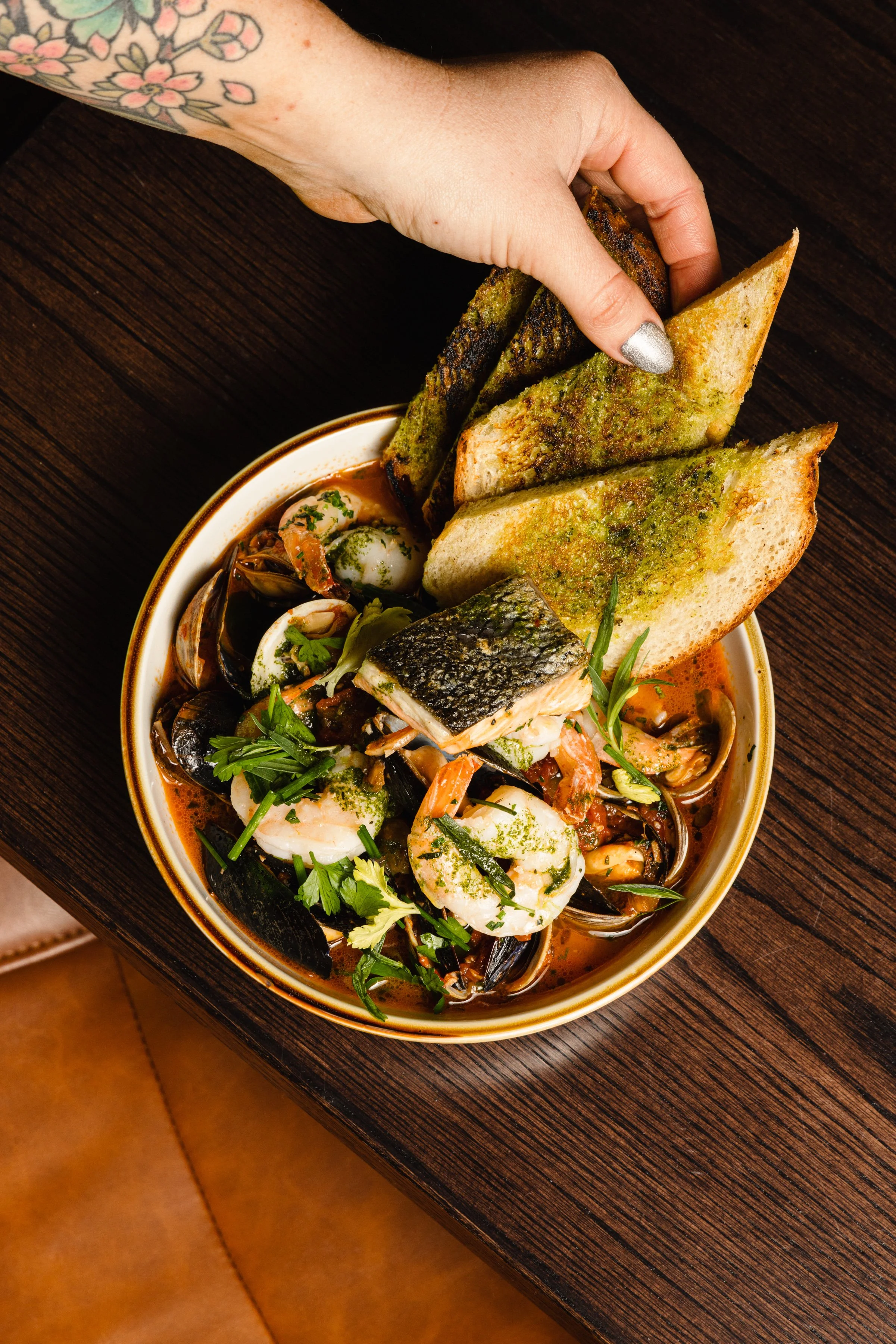 Hand with a tattoo reaching over a bowl of seafood including shrimp, fish, and mussels, with toasted bread slices on top, on a dark wooden table.