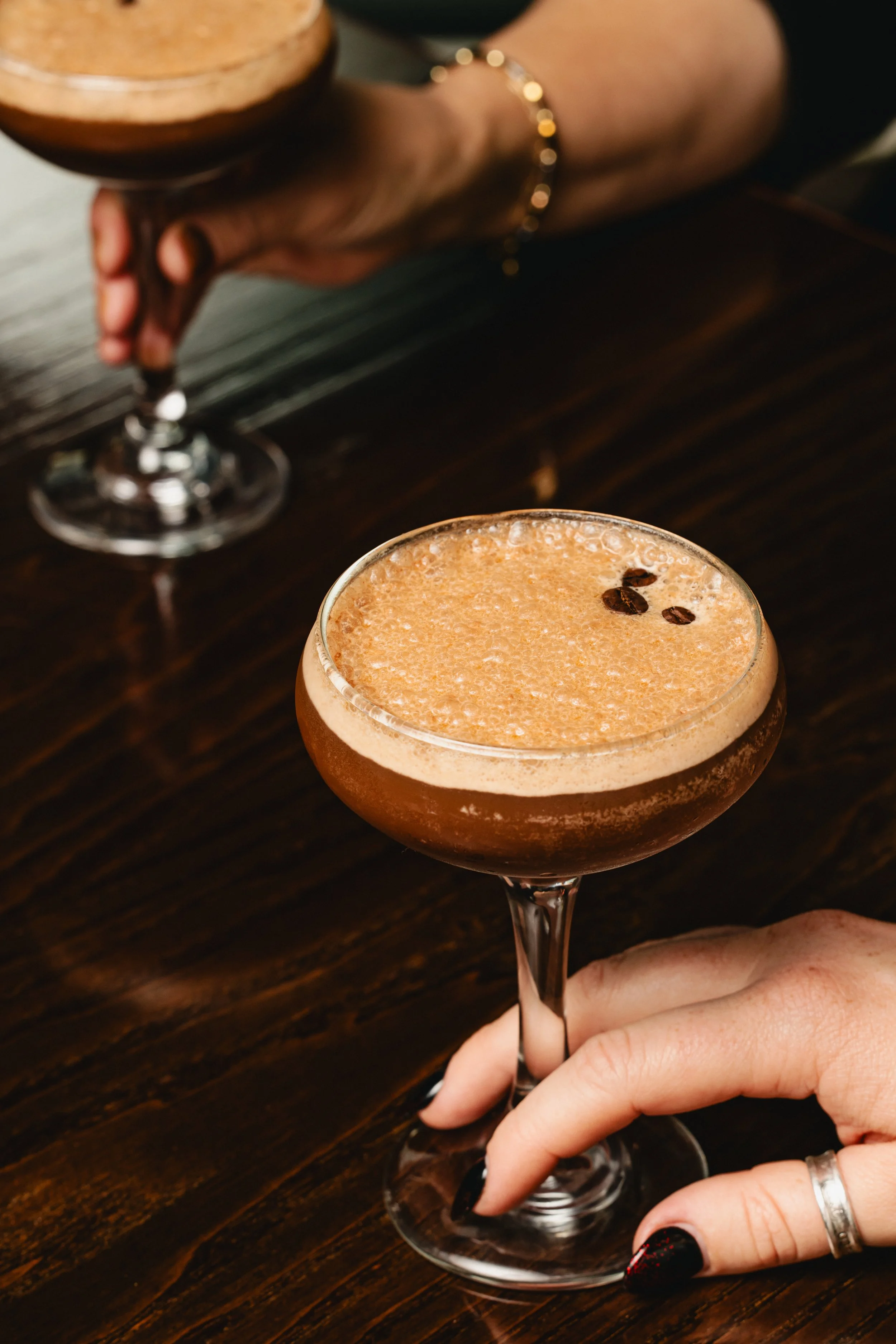 Close-up of a cocktail in a coupe glass with coffee beans garnishing, held by a woman with dark nail polish and a silver ring, on a dark wooden table.