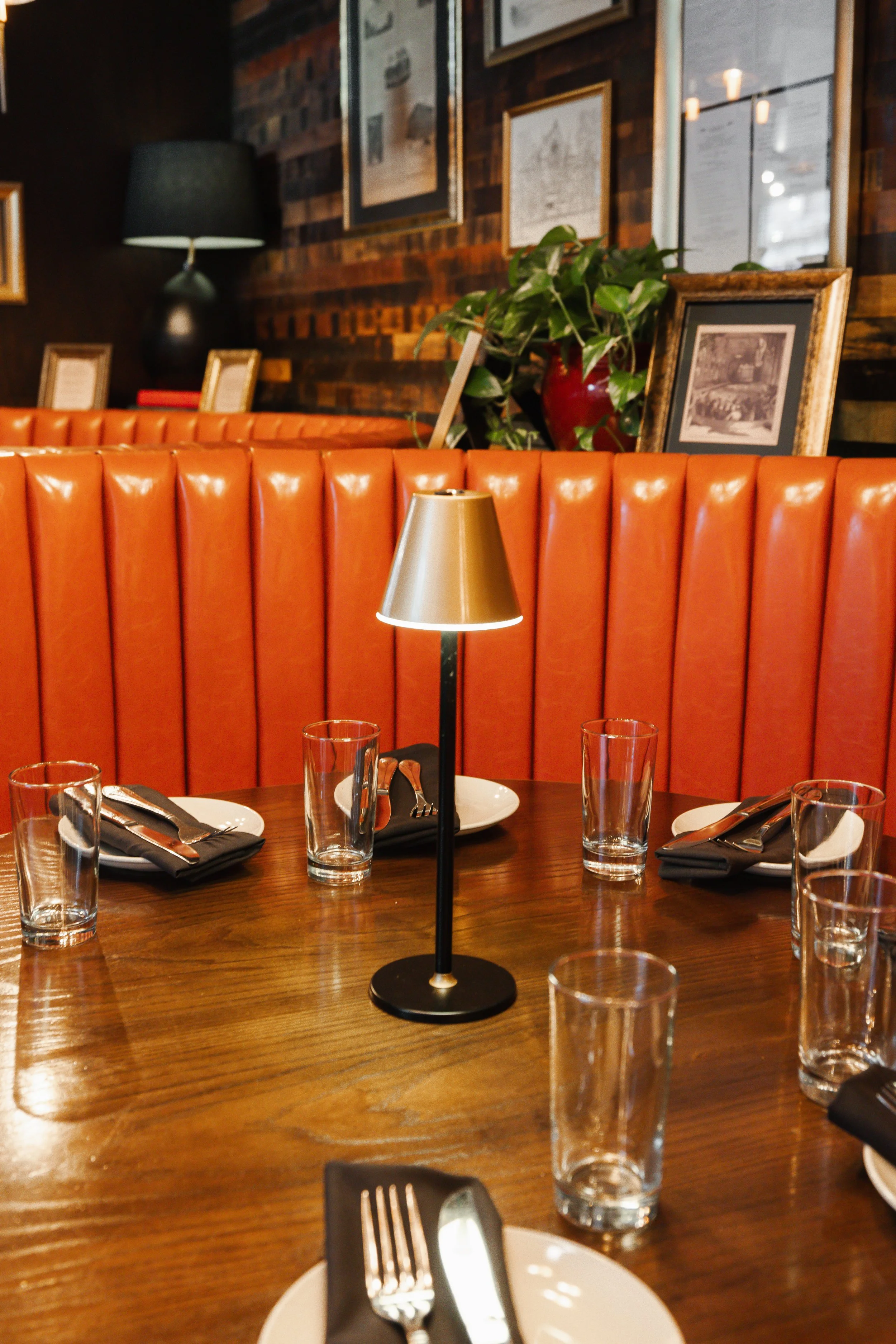 A round wooden dining table set for a meal with black napkins, silverware, and empty glasses, featuring a small black and gold table lamp in the center, with a red booth, framed pictures, green plant, and wall decor in a cozy dimly lit restaurant or