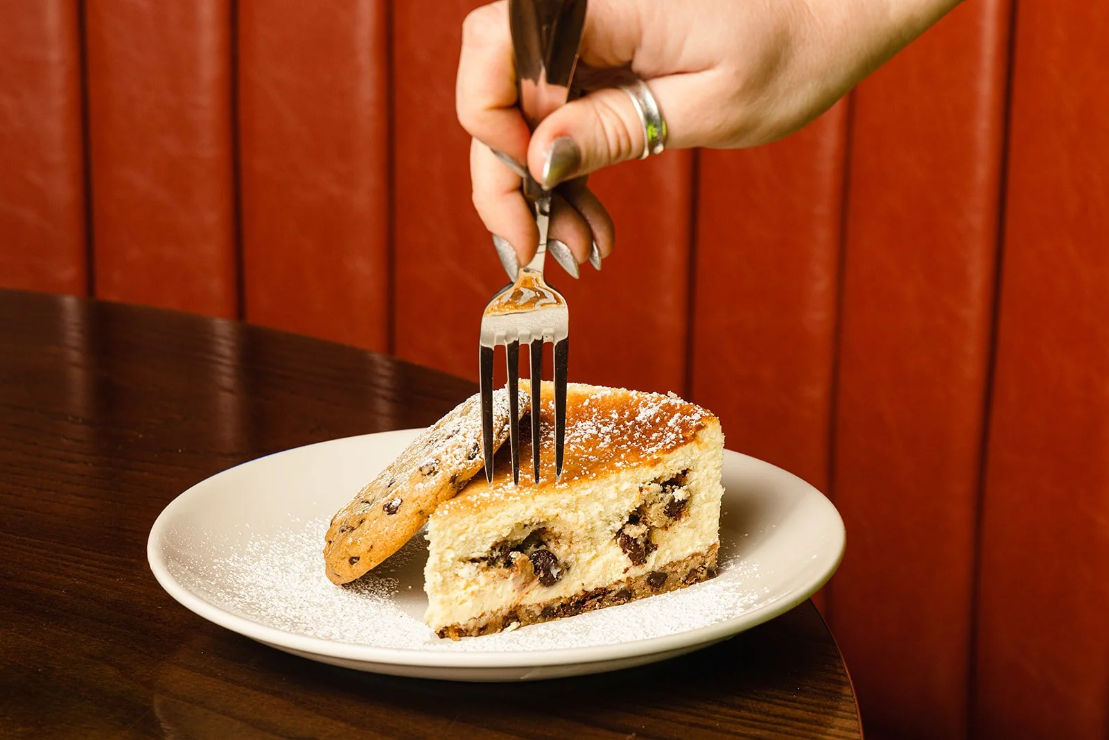 A hand with painted nails and rings is holding a fork, about to take a bite of a slice of cheesecake with chocolate chips, on a white plate dusted with powdered sugar, with a chocolate chip cookie leaning against it, on a dark wooden table with red u