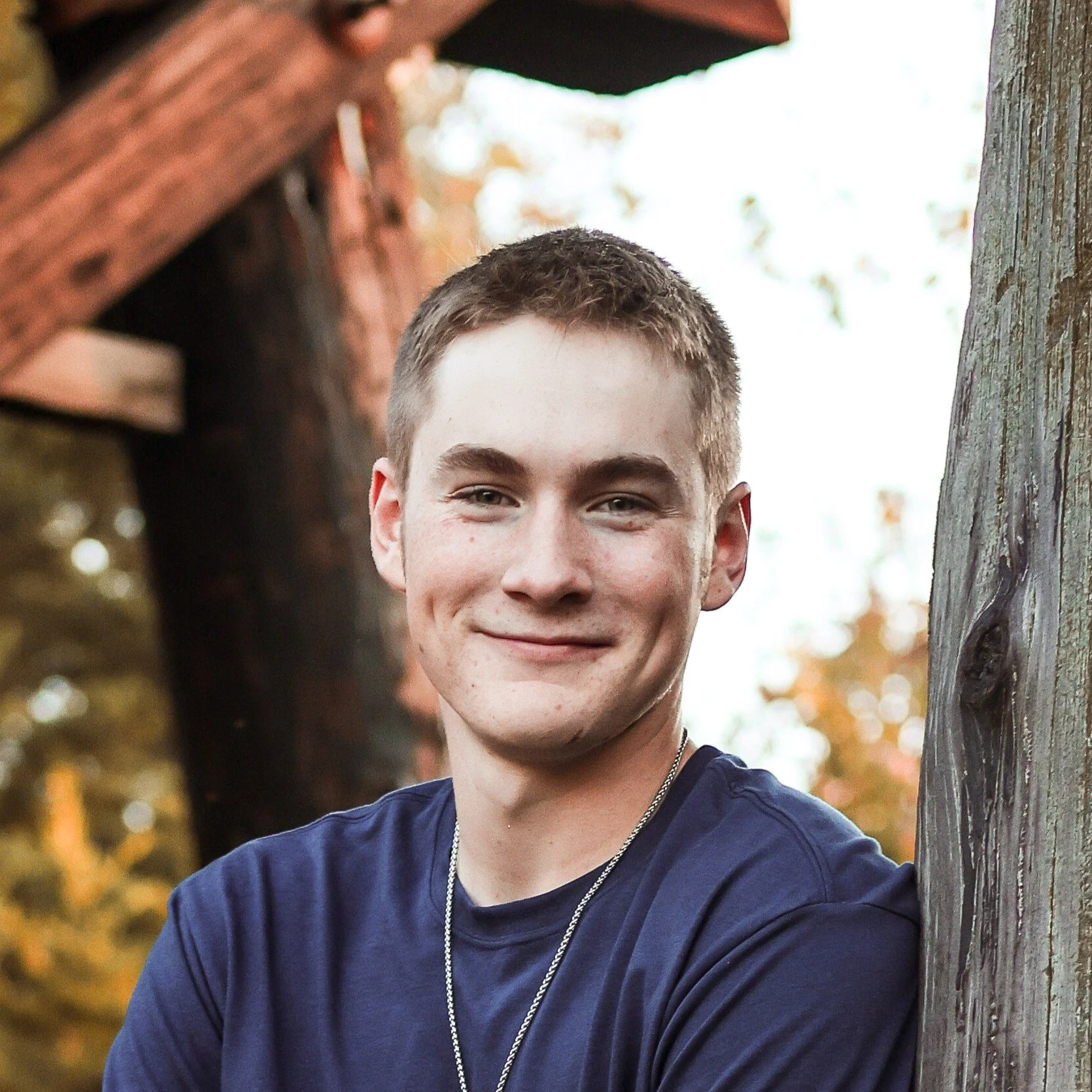 Young man with short hair wearing a navy blue shirt and silver chain necklace outdoors during autumn, leaning against a weathered wooden post with blurred fall foliage in the background.