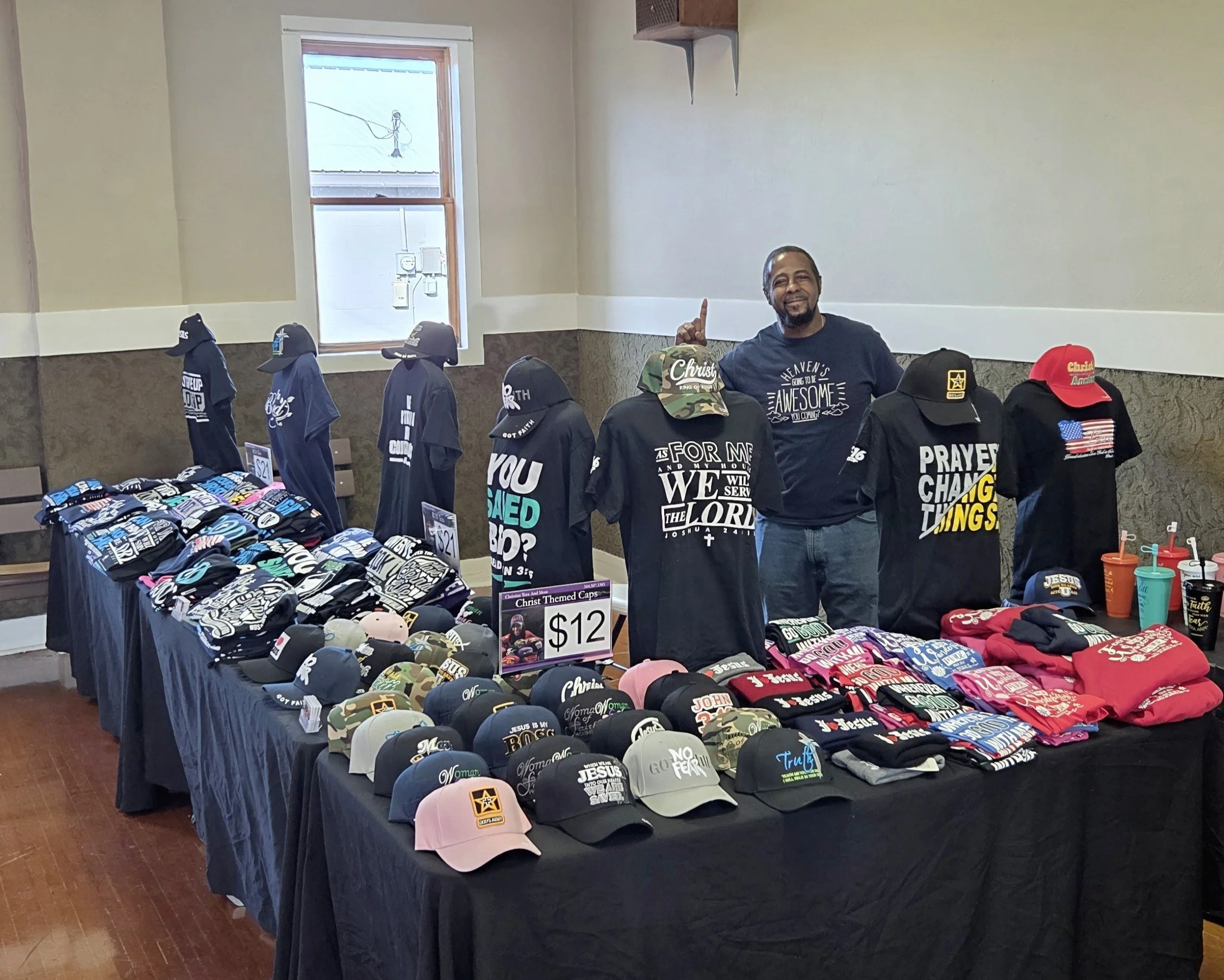 A man standing behind a display table of Christ-themed hats and t-shirts at a store or market. The table has various hats with religious messages and graphics, and t-shirts with related text. The man is smiling and pointing upward with his finger. The background shows a window and part of the indoor space.