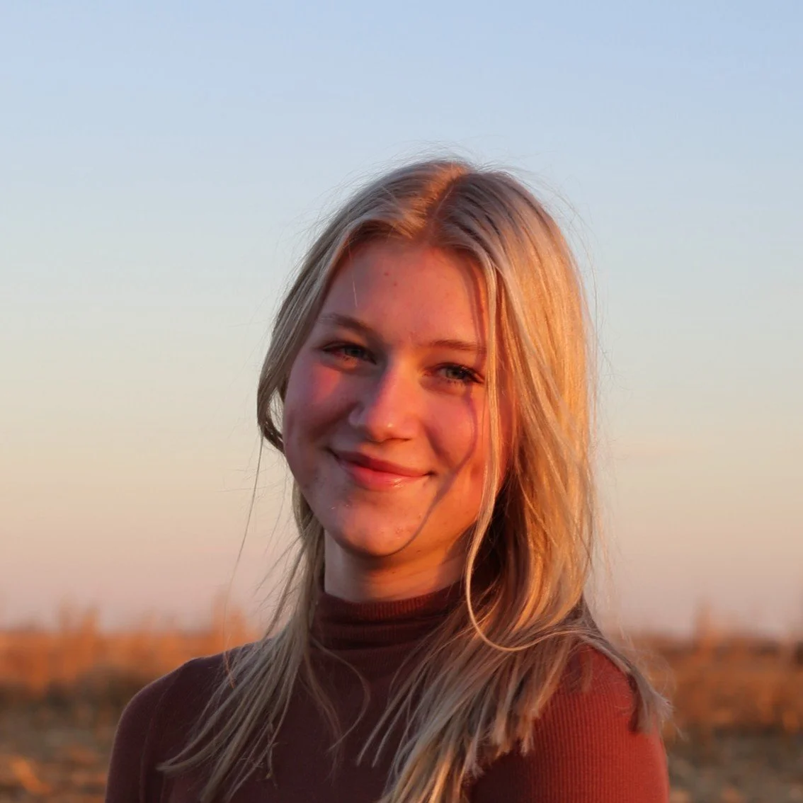 A young woman smiling outdoors during sunset, with a clear sky in the background.