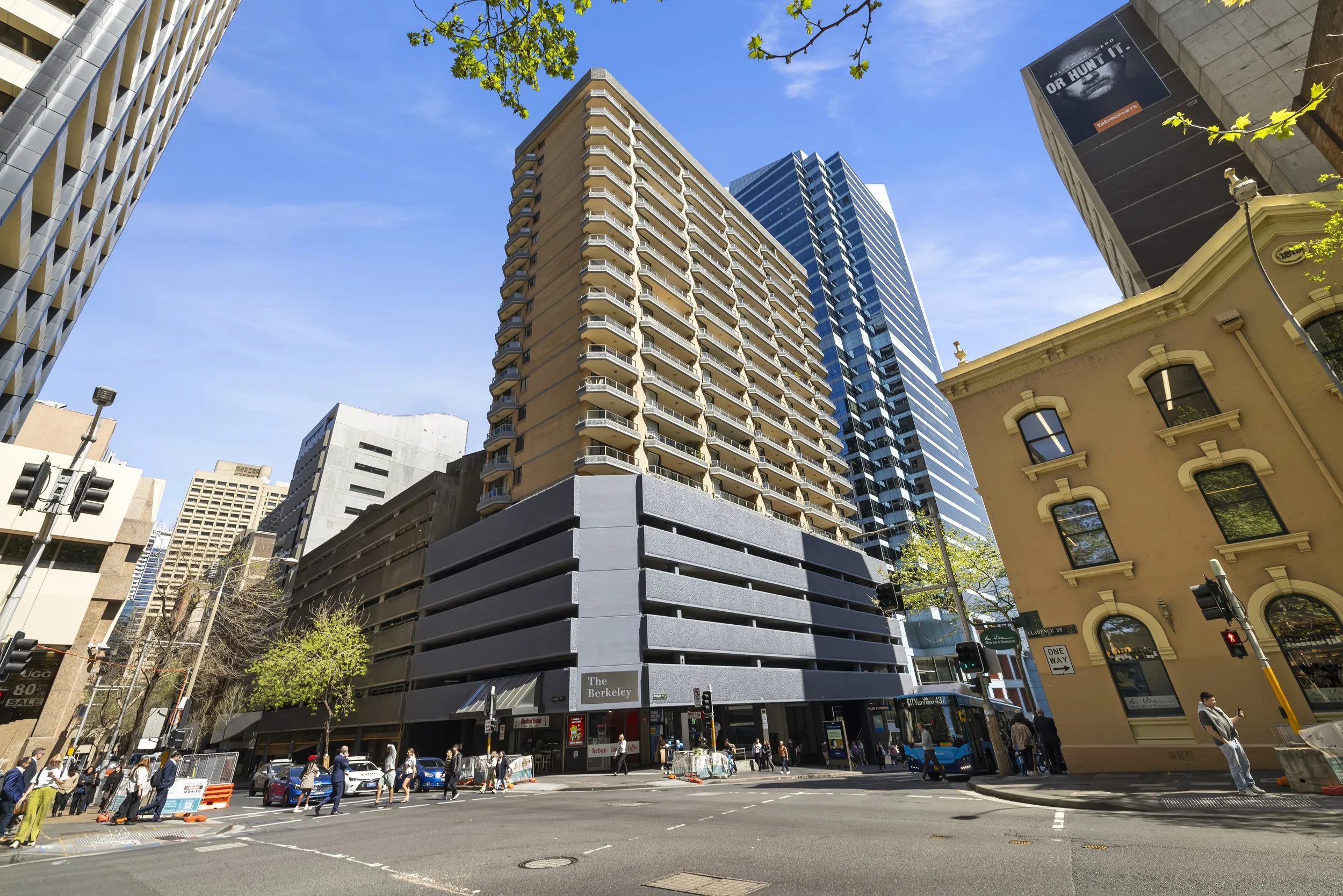A city street scene with a tall modern building at the corner, people waiting at a crosswalk, a bus on the street, and other high-rise buildings in the background under a blue sky.
