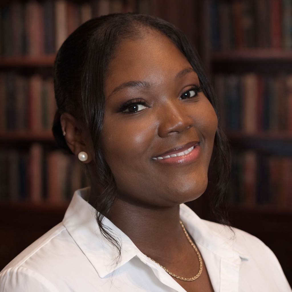 A woman with dark hair styled in loose waves, wearing pearl earrings, a white collared shirt, and a gold chain necklace, smiling with a background of bookshelves filled with books.