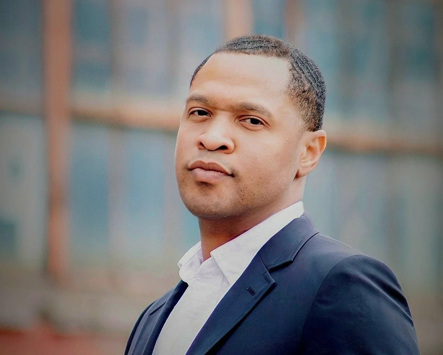 A man with short dark hair, wearing a dark suit jacket and a white shirt, looking at the camera with a neutral expression, outdoors with blurry background.