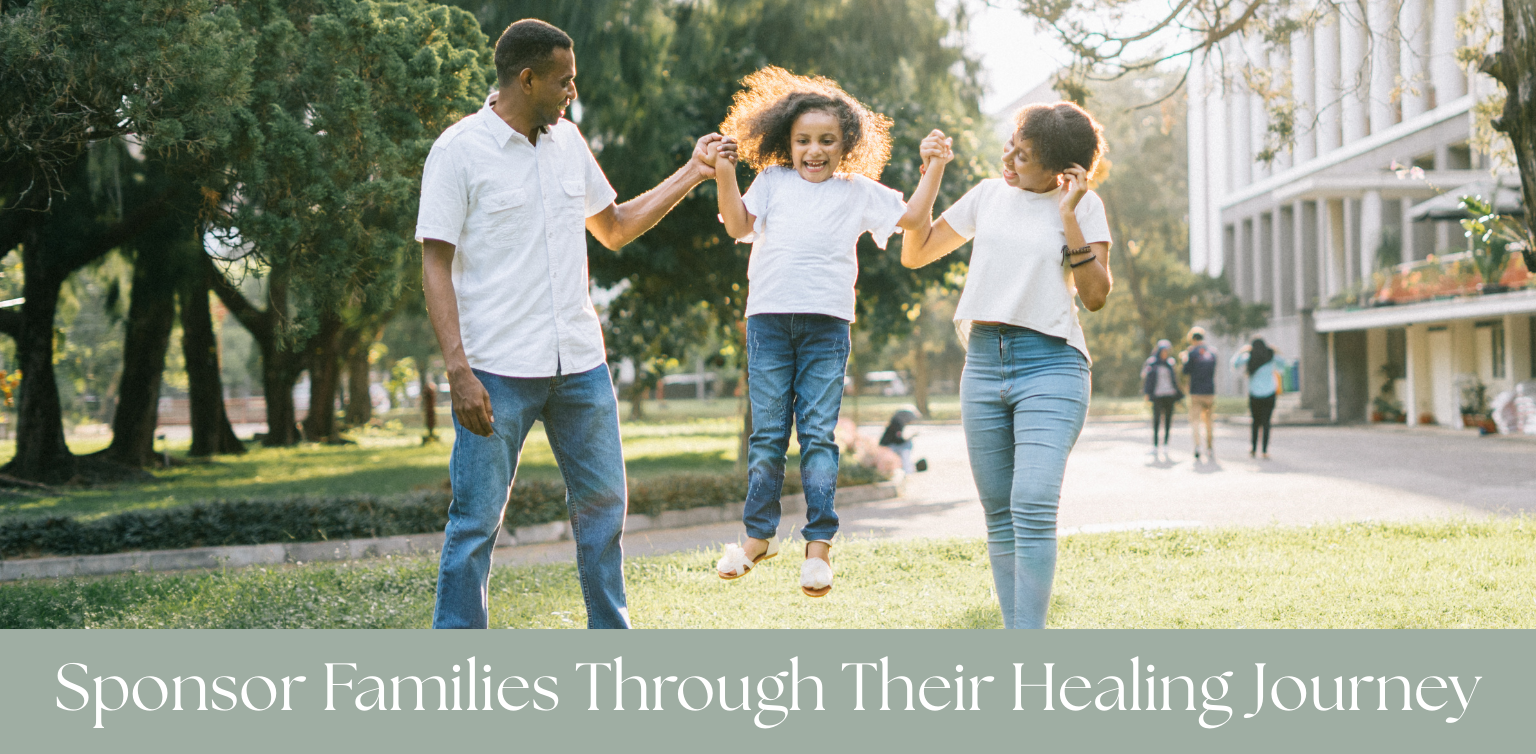 A family of three, two adults and a young girl, enjoying outdoor time in a park. The girl is jumping between her parents, who are holding her hands, with trees and other park visitors in the background.