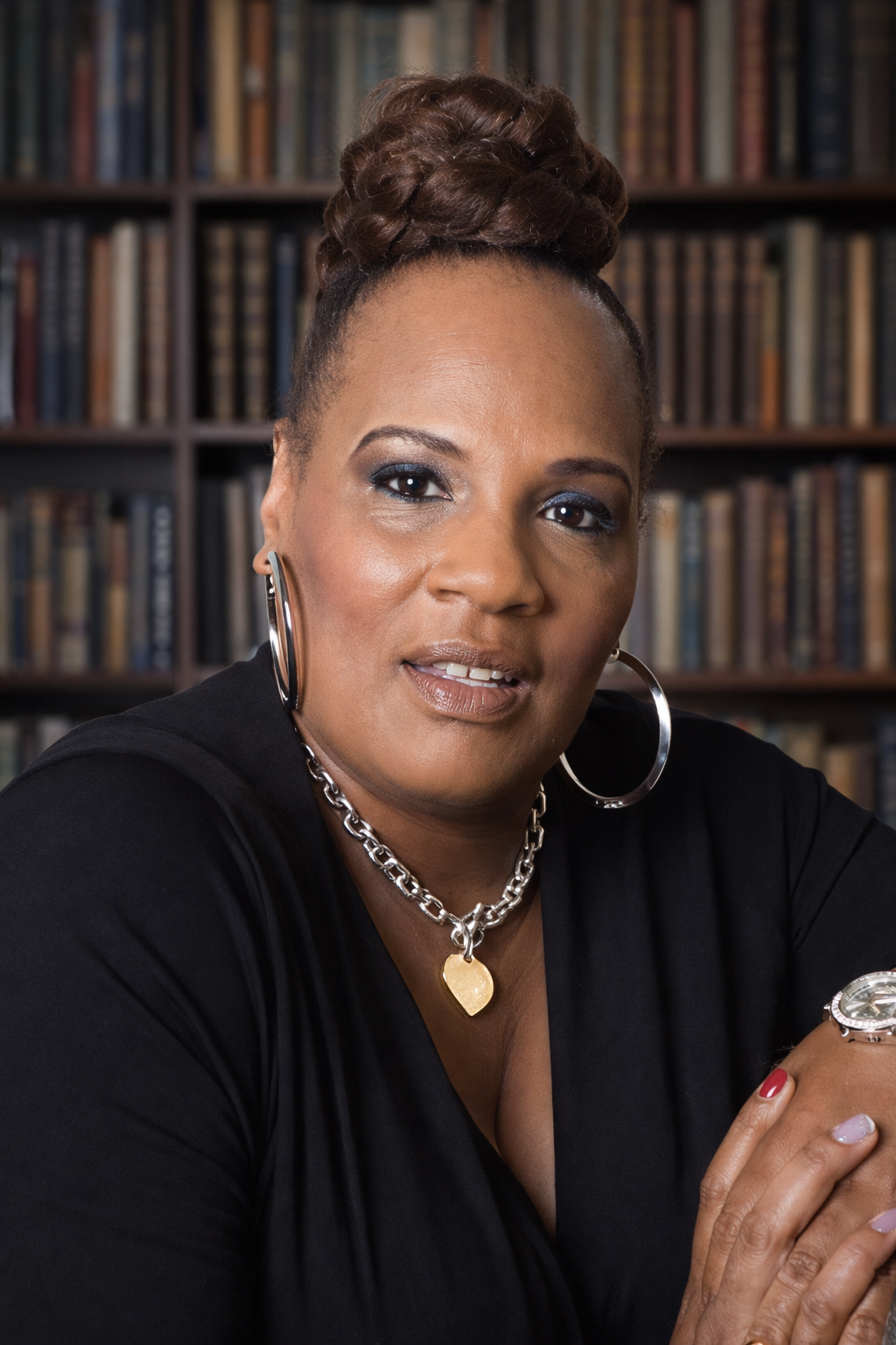 A woman with dark skin, curly hair in an updo, wearing large hoop earrings, a silver chain necklace with a gold heart pendant, and a black top, seated in front of a bookshelf.